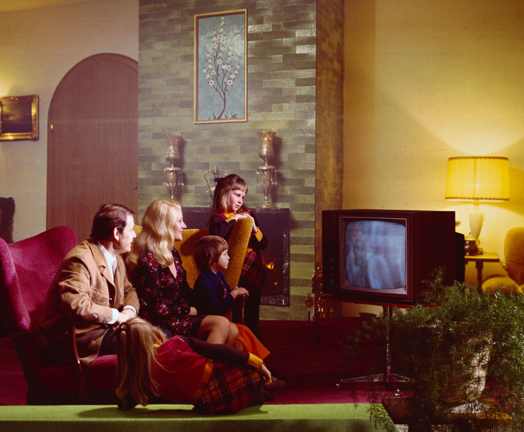 A family of five, including a man, a woman, two girls, and a boy, sits on a couch and floor in a living room, watching a television together