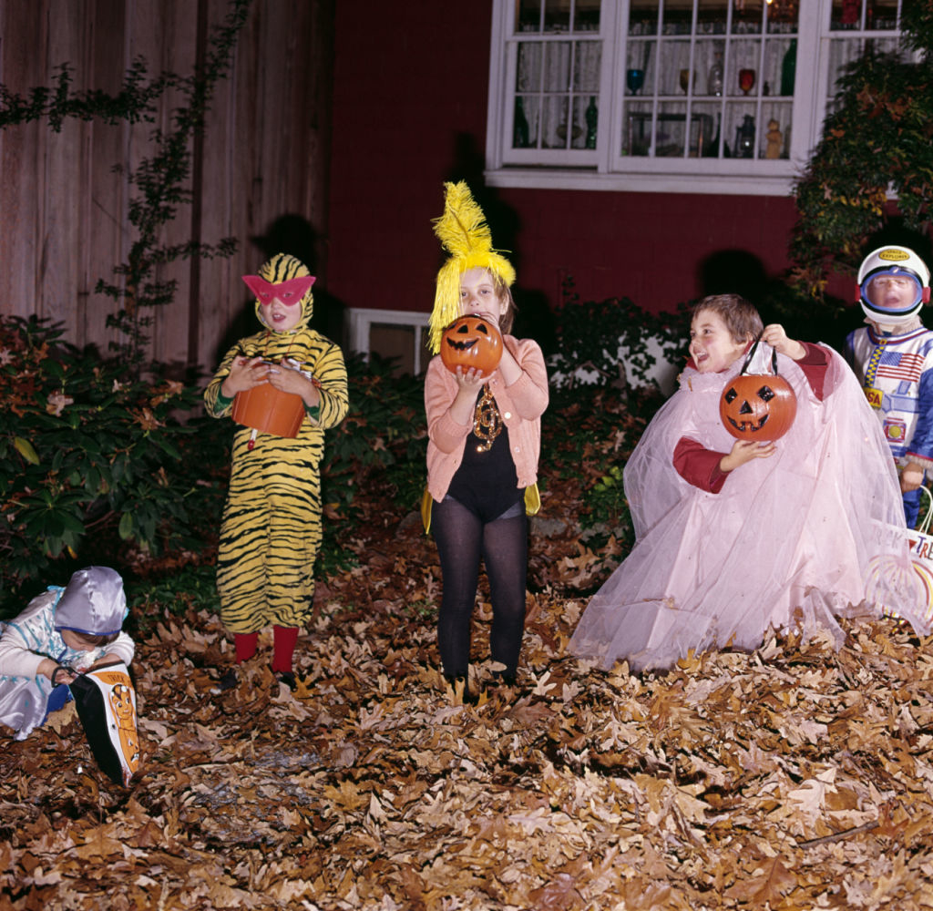 A group of children dressed in various Halloween costumes, holding pumpkin buckets, posing in front of a house