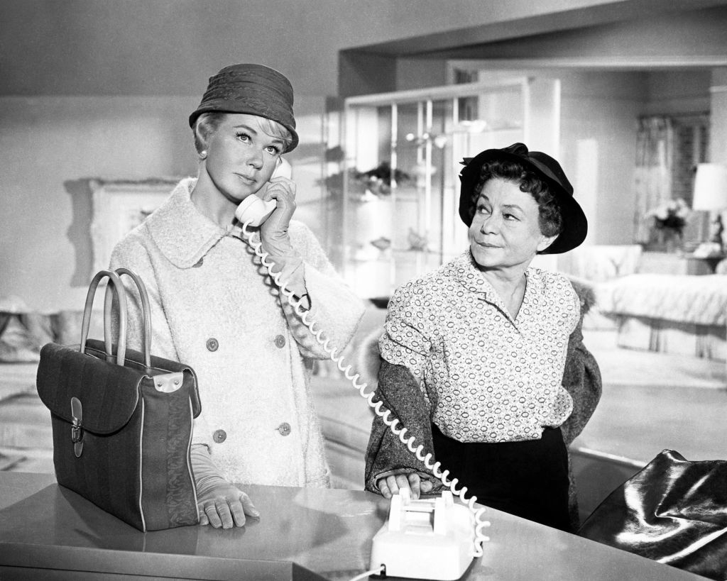 Doris Day, wearing a coat and hat, speaks on a landline phone, while Thelma Ritter, in a patterned blouse and hat, stands beside her in a modern office setting in a scene from the movie Pillow Talk