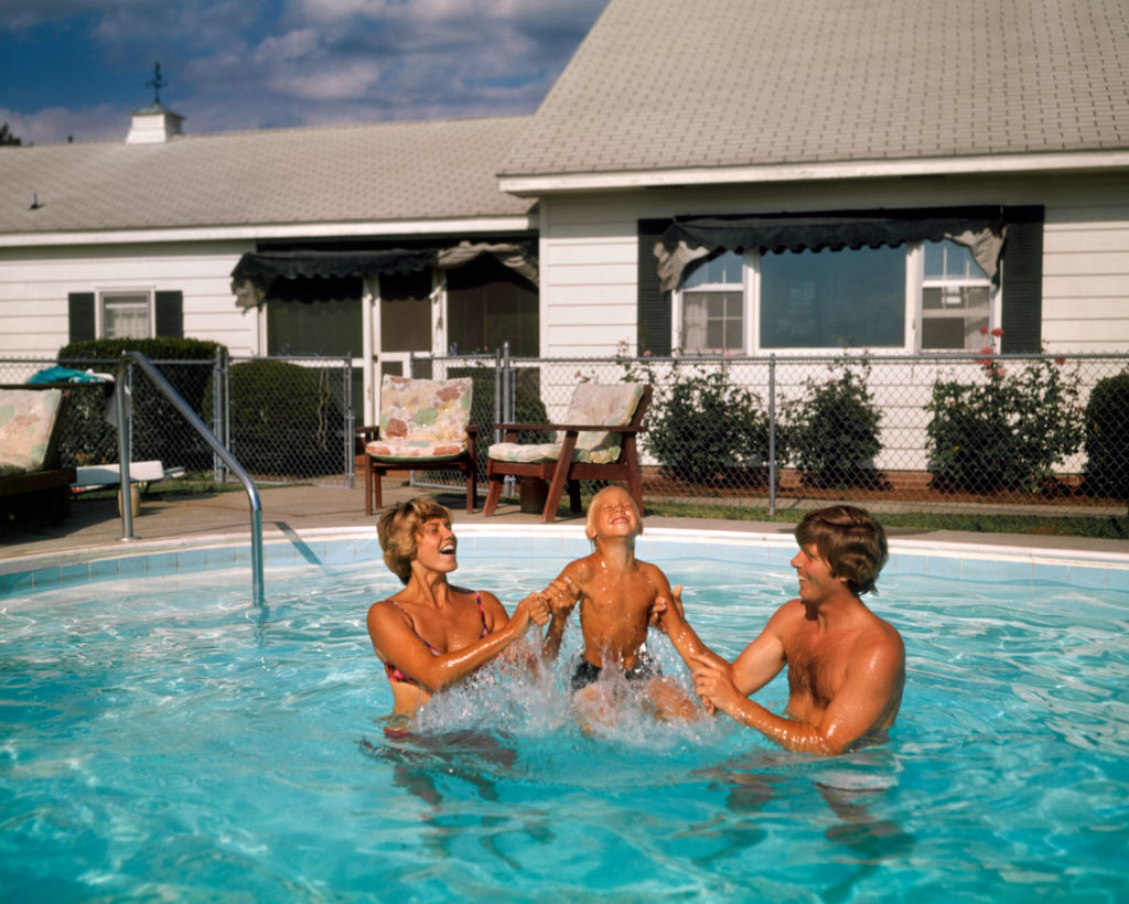 A man and a woman in a pool play with a young boy at their suburban home