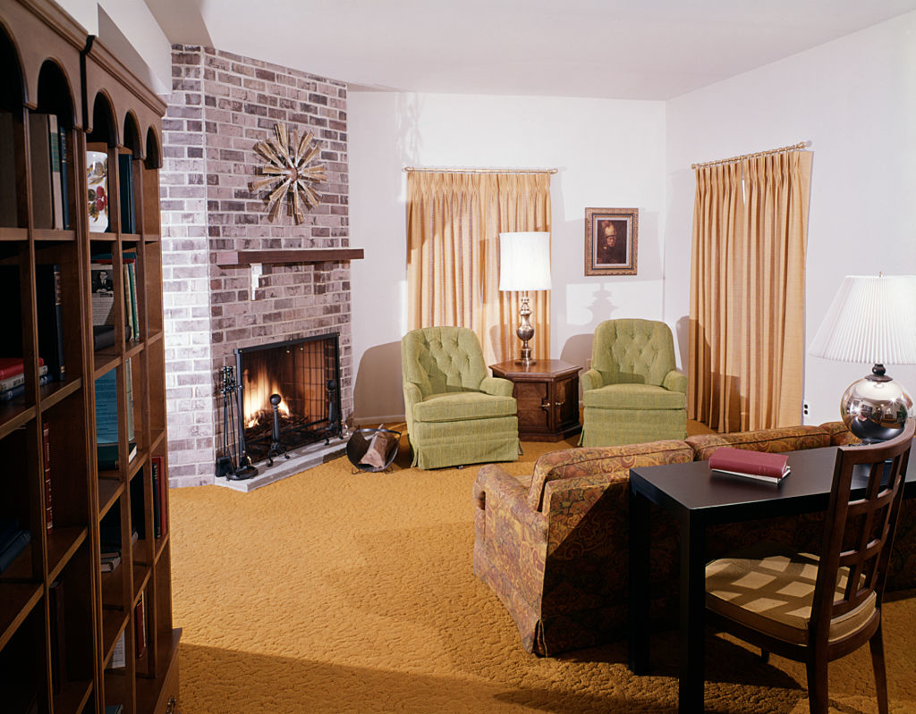 Living room with brick fireplace, green chairs, a wooden side table, a couch, and a black desk with a lamp and book. Picture of a woman on the wall