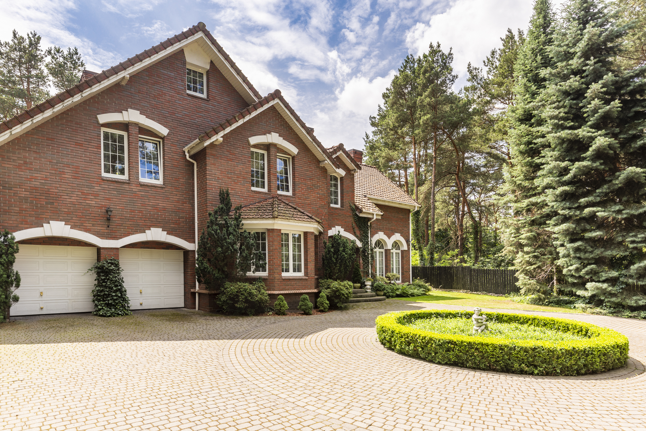 A spacious brick house with a three-car garage, surrounded by trees and neatly landscaped with a circular hedge design in the driveway