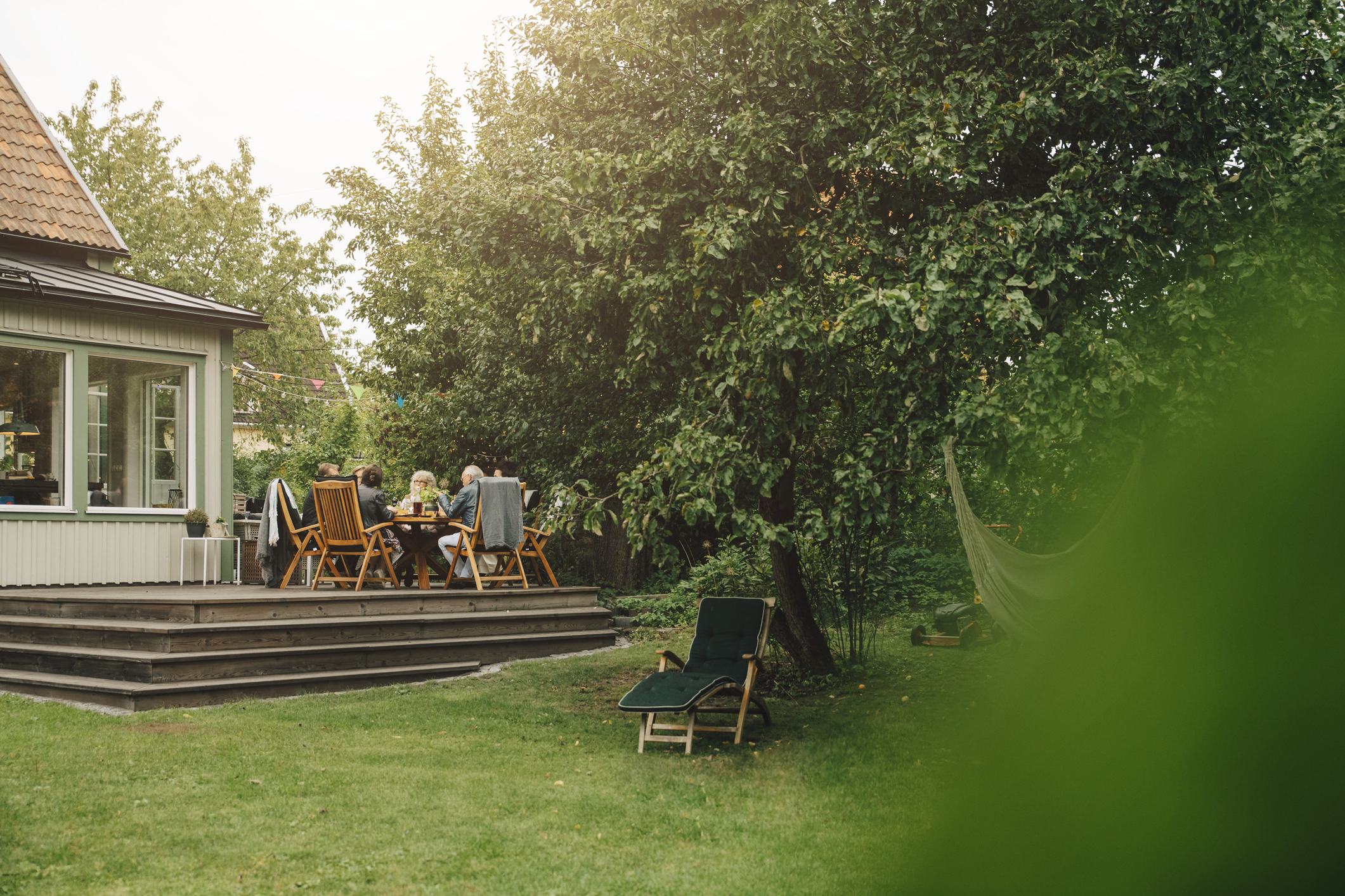 People are seated at a wooden table on a backyard deck next to a house, surrounded by trees and greenery