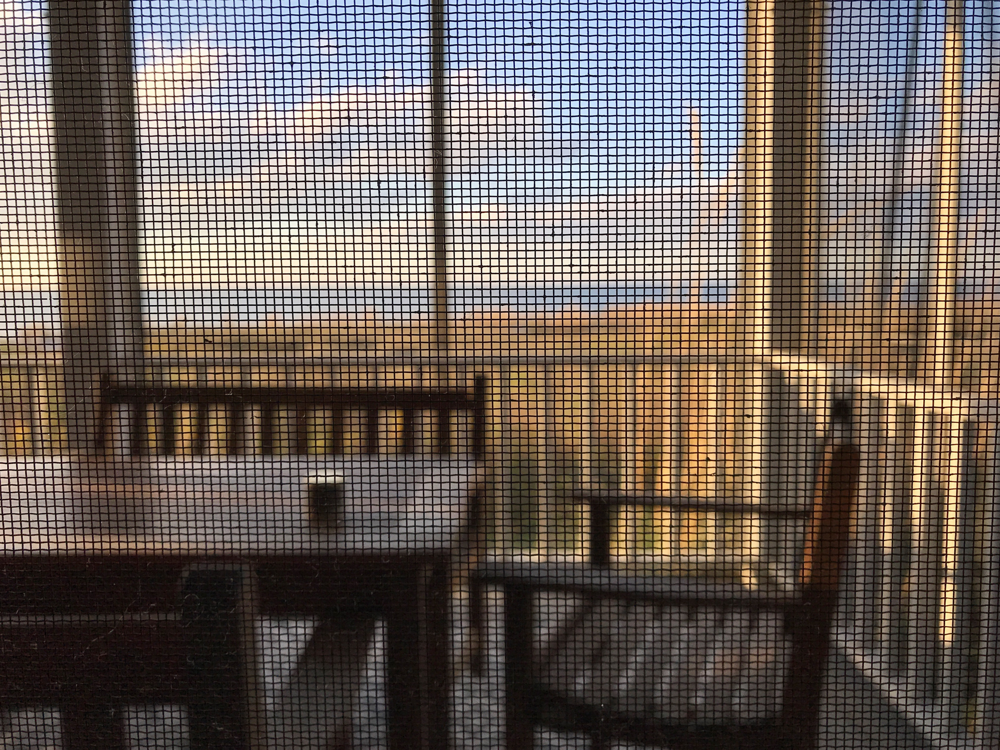 View of an outdoor seating area with wooden furniture, seen through a mesh screen, overlooking a scenic landscape. No people are in the image