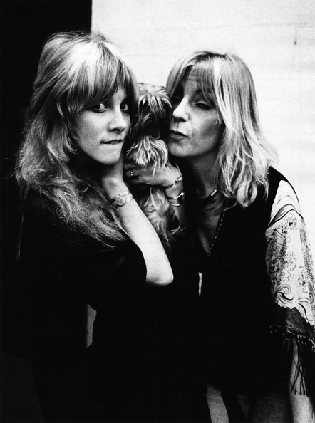 Stevie Nicks and Christine McVie pose while holding a small dog, smiling and in casual clothing