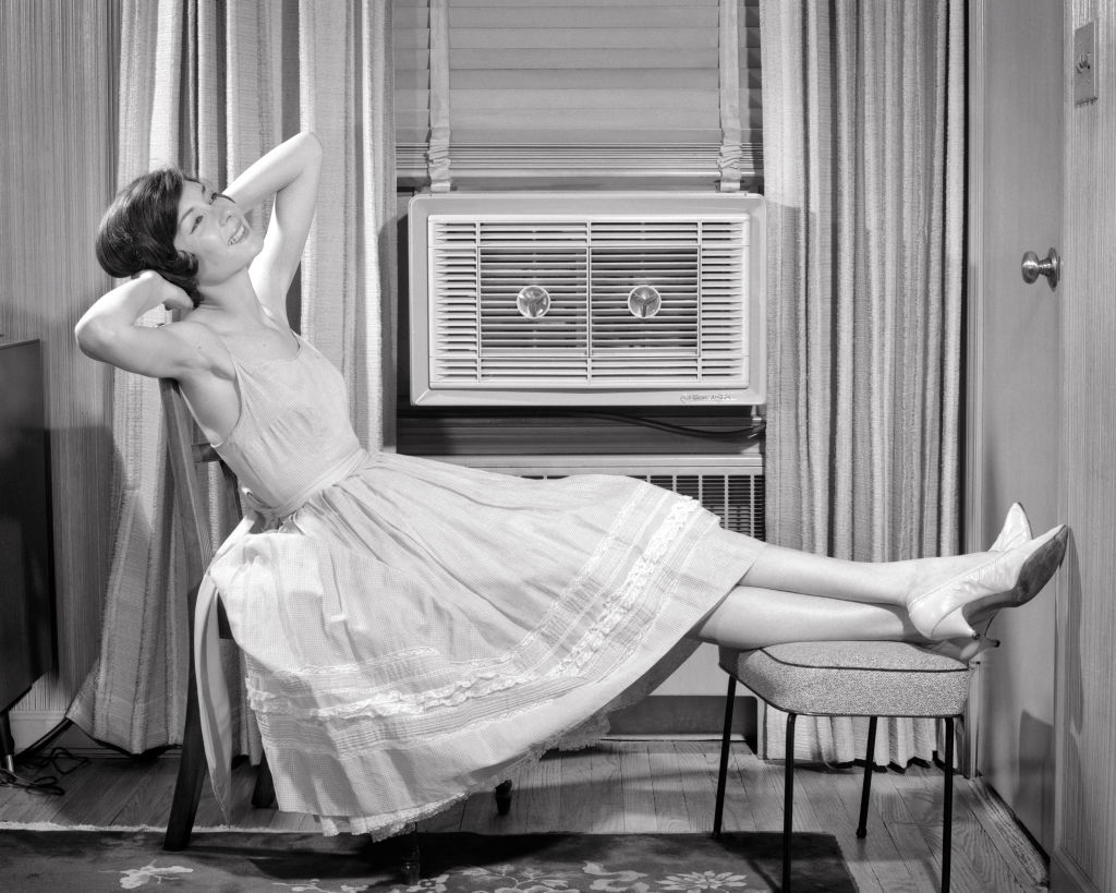 A woman in a sleeveless dress lounges on a chair with her feet propped up on a small stool, smiling and appearing relaxed in a well-appointed living room