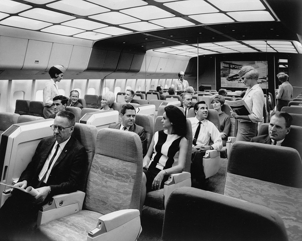 Passengers are seated in a vintage airplane cabin. Flight attendants are serving and assisting passengers. The cabin features spacious seating and overhead lighting