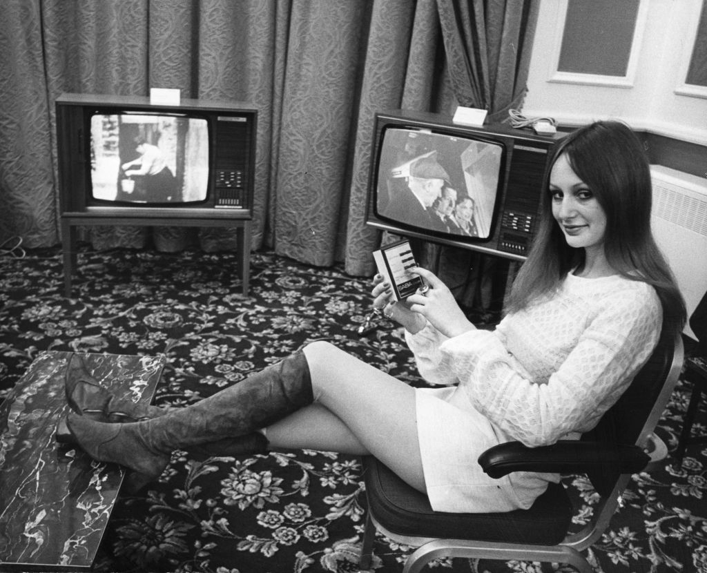 A woman in a short dress and knee-high boots sits by two vintage televisions in a retro-styled room, holding a small object
