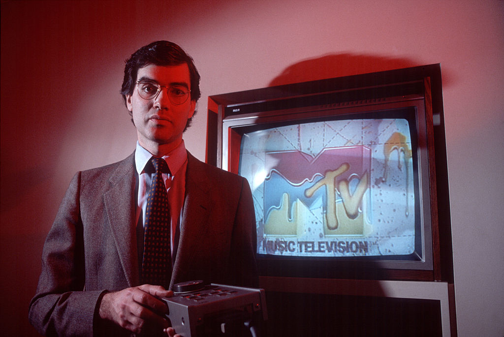 A man in a suit and tie stands holding a device in front of an early MTV television displaying the channel’s logo