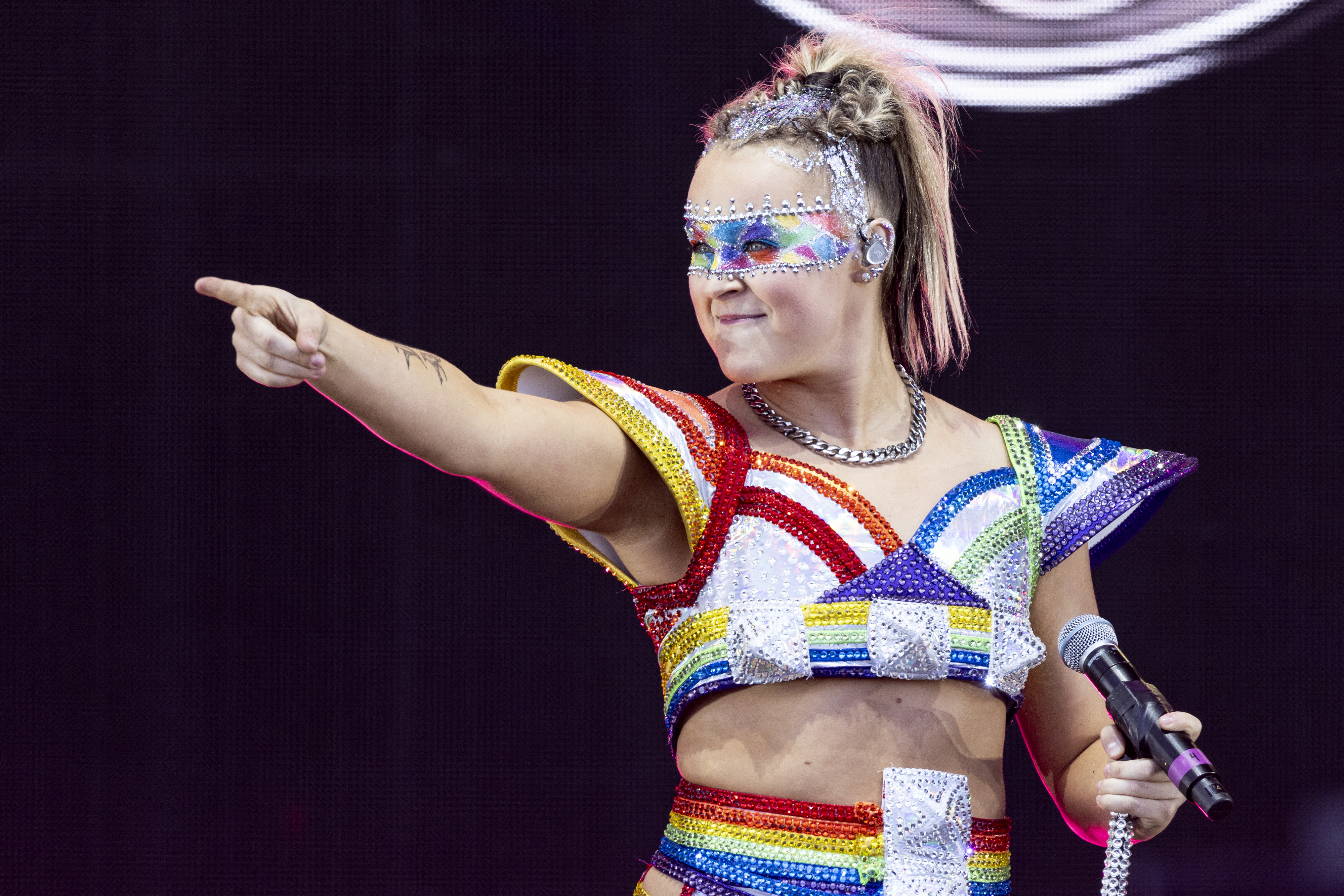 JoJo Siwa performing on stage in a sparkling, rainbow-themed costume with geometric designs, holding a microphone and pointing outward