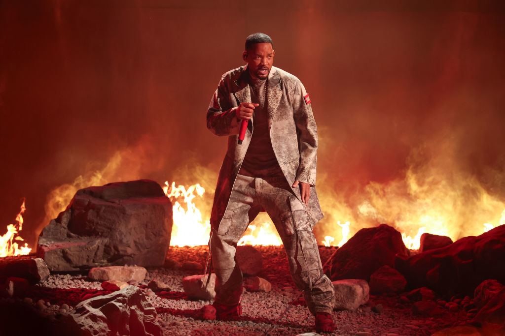 Will Smith performs on stage in military-style clothing, with a fiery background and rocks scattered on the ground