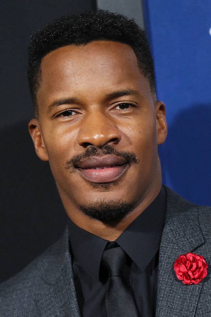 Nate Parker wears a dark suit, black shirt, and black tie with a red flower lapel pin, smiling at a formal event