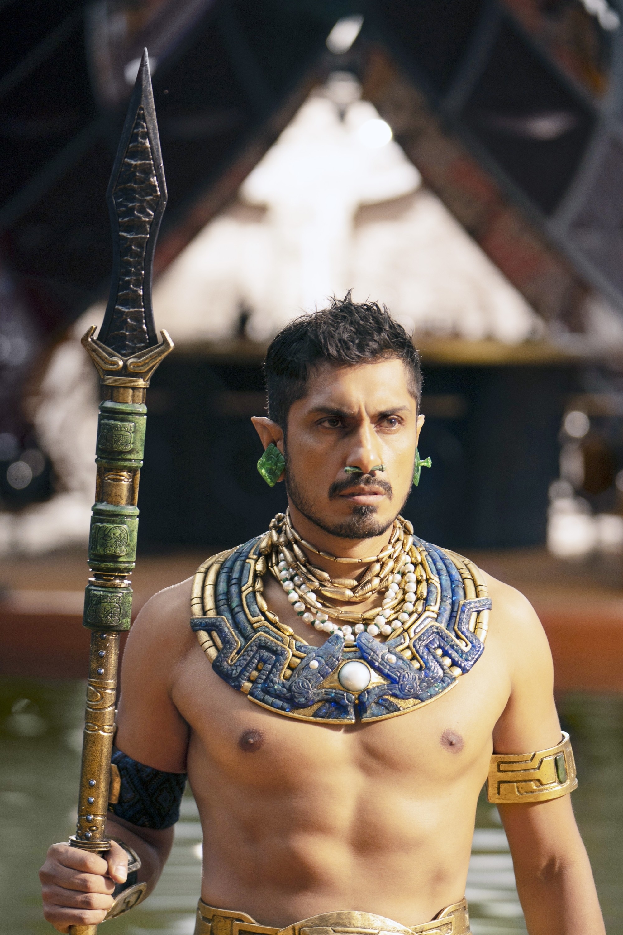 Tenoch Huerta, shirtless in a warrior role, holding a spear, wearing elaborate gold and turquoise accessories, looking intently ahead