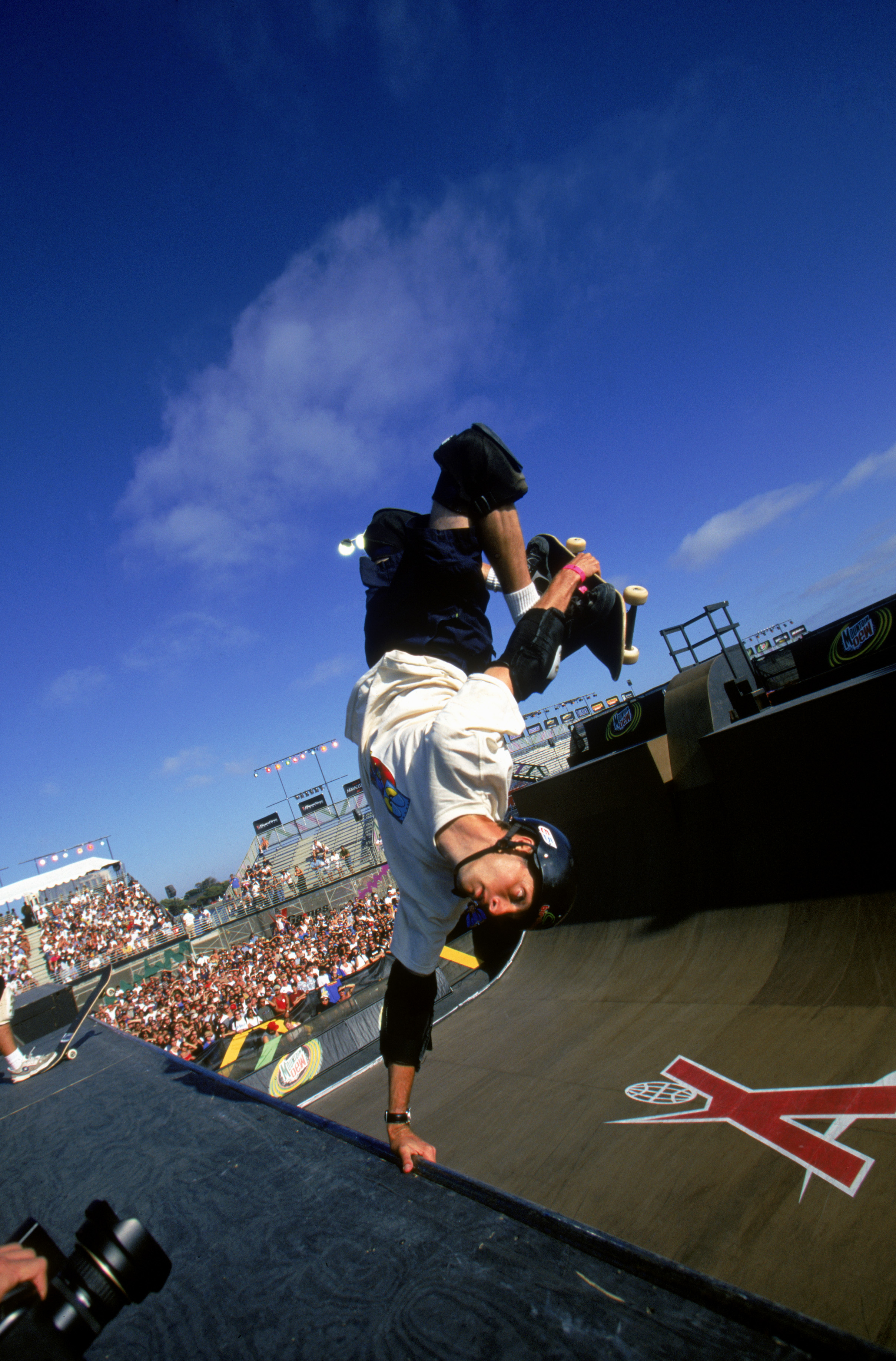 Tony Hawk performs an upside-down skateboarding trick on a ramp at a skateboarding event, with spectators in the background