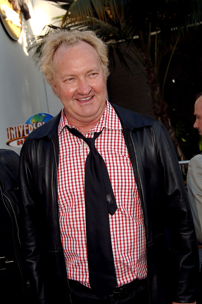 A man with blond hair wearing a red and white checkered shirt, black tie, and black leather jacket at an outdoor event