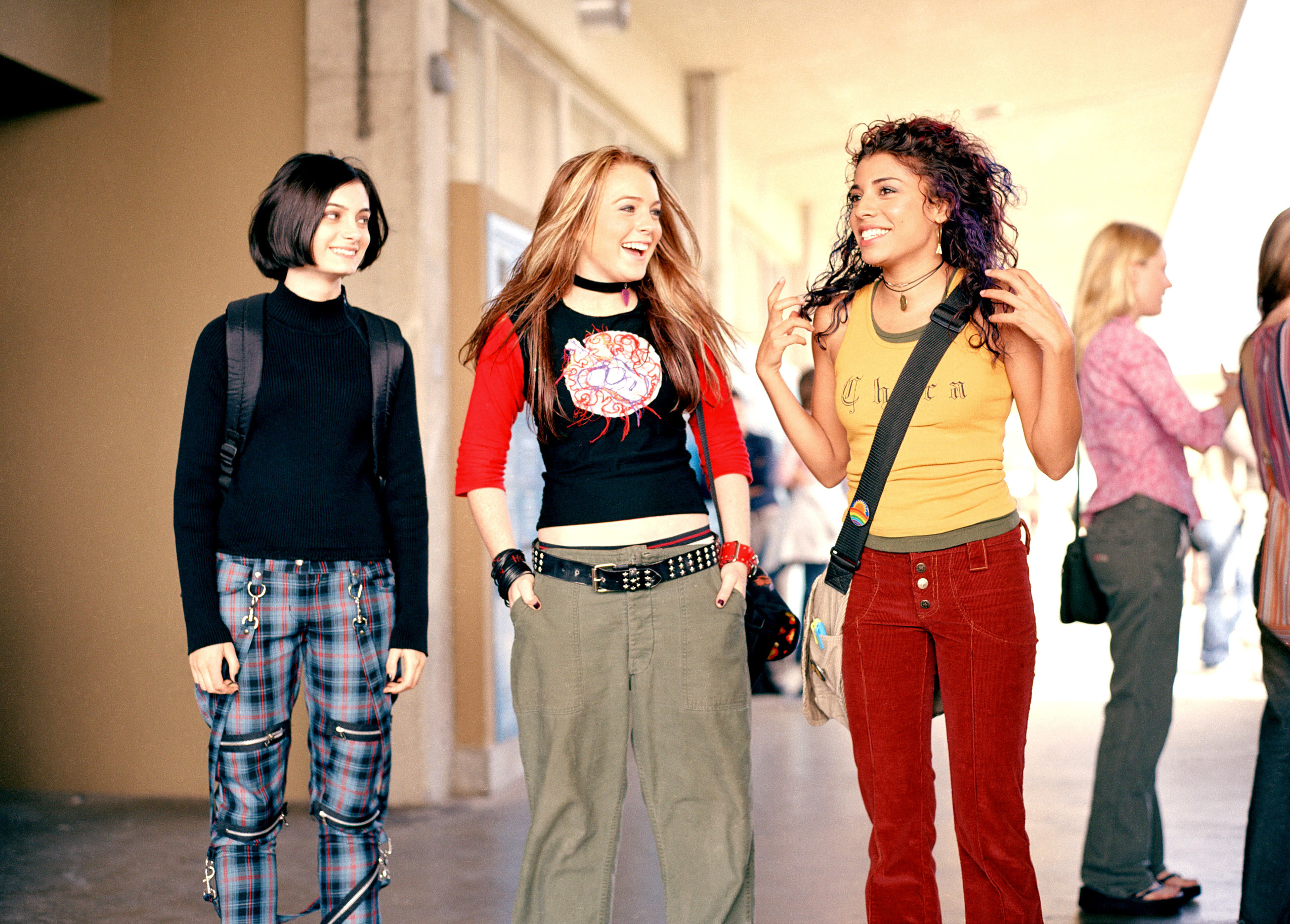 Lindsay Lohan, wearing casual pants and a KISS logo shirt, walks with two friends in casual outfits, smiling and chatting in a hallway in a scene from Freaky Friday