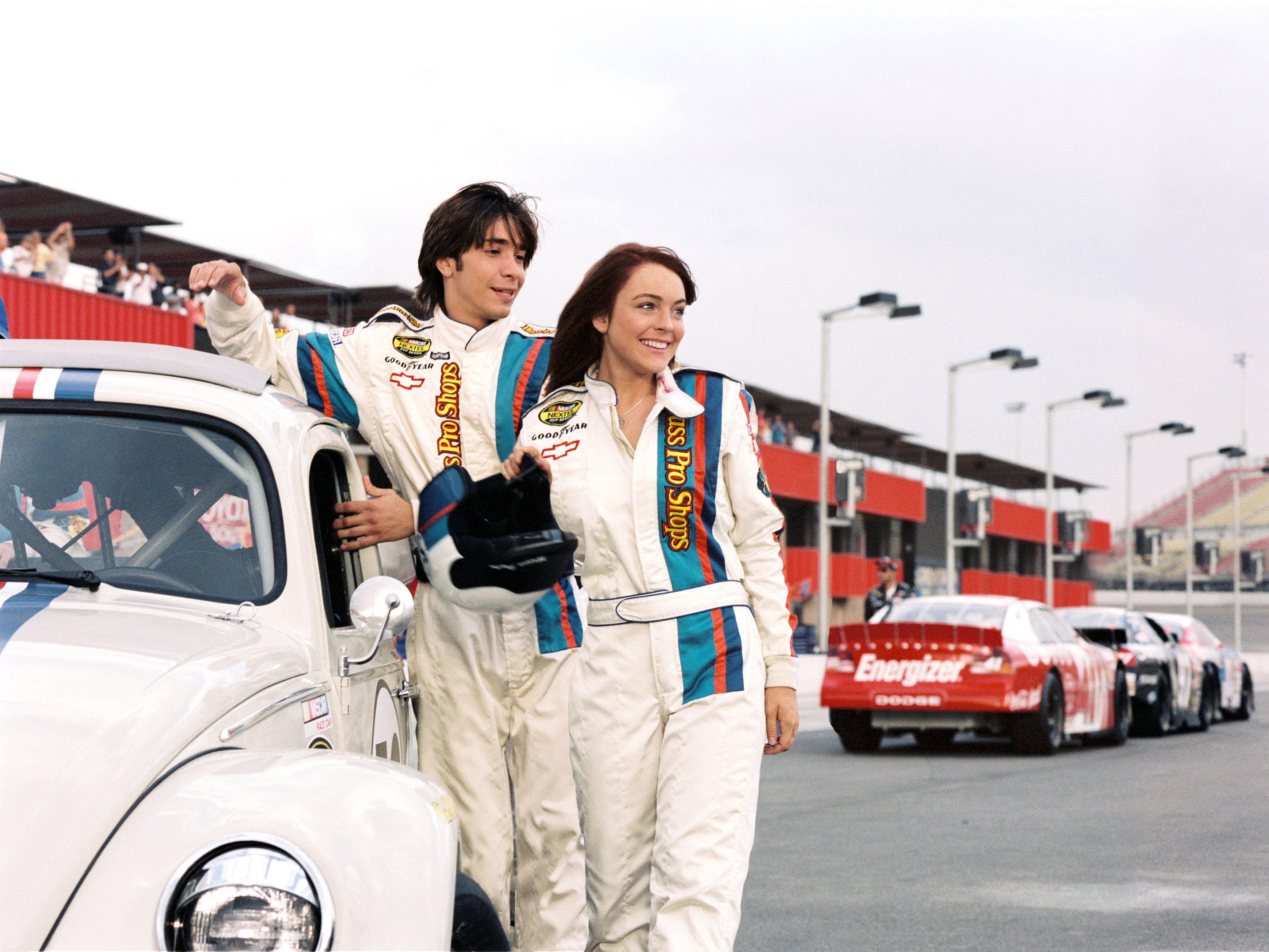 Lindsay Lohan and Justin Long in racing suits standing next to a vintage car on a racetrack with race cars in the background