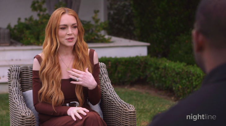 Lindsay Lohan in a seated outdoor interview, wearing a dress with shoulder cut-outs, gesturing with her hand, green foliage in the background