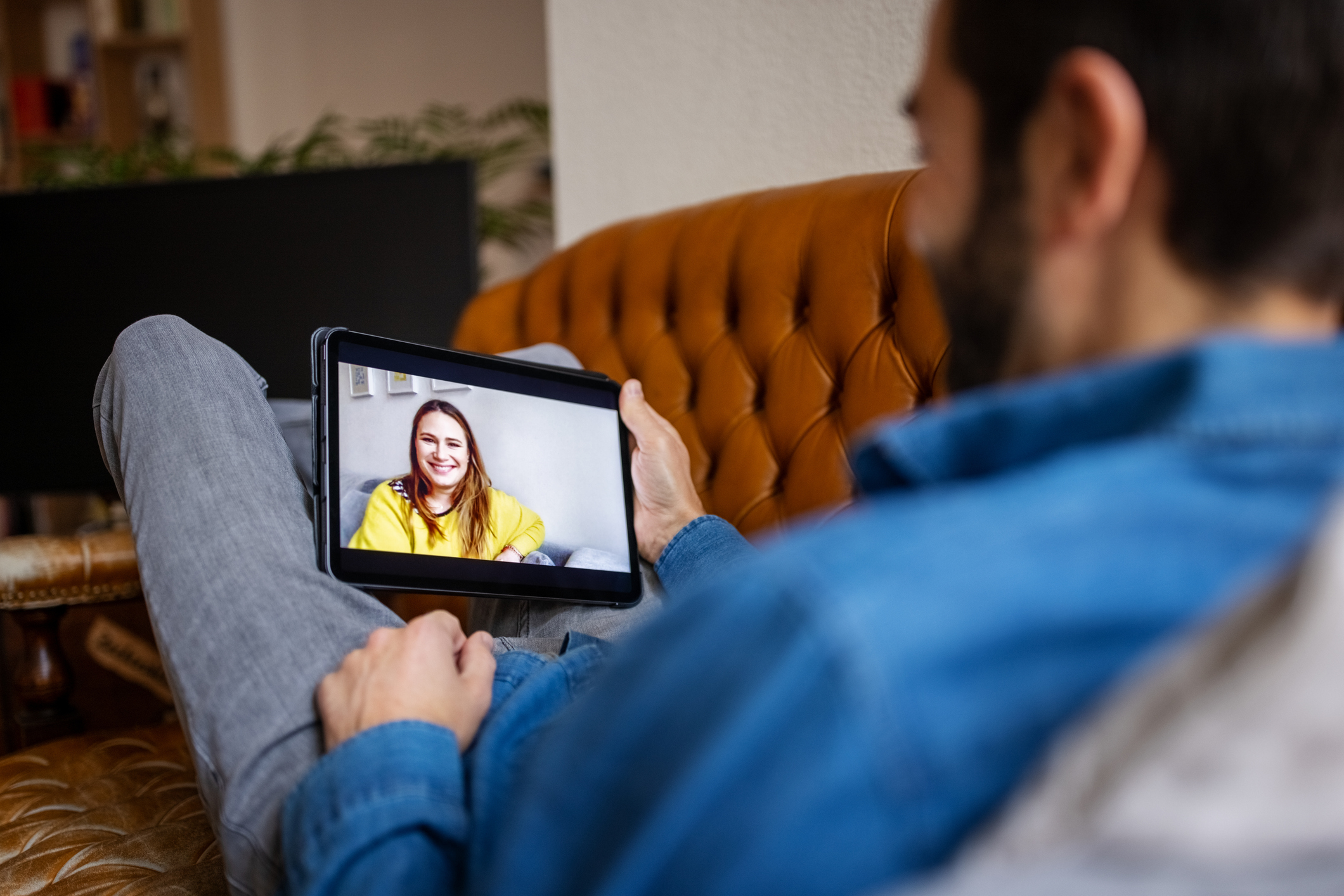 A person is reclining on a couch, holding a tablet showing a video call with a smiling woman wearing a casual top