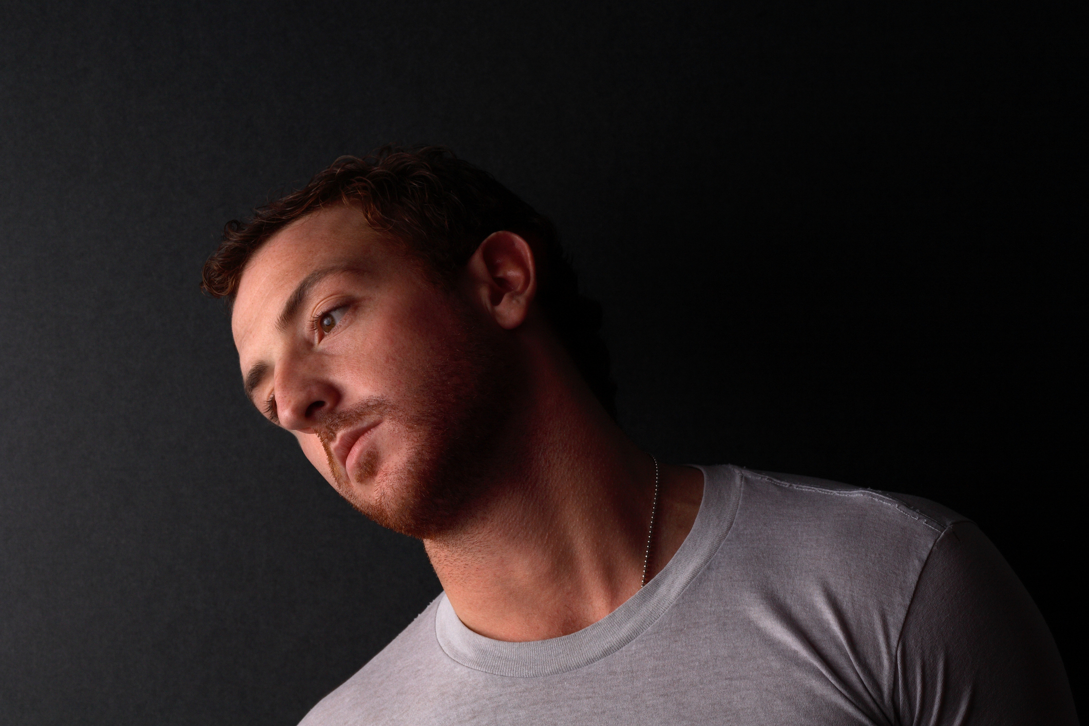 A man with short hair and a trimmed beard tilts his head to the side against a dark background. He is wearing a plain T-shirt. Names of people in the image are unknown