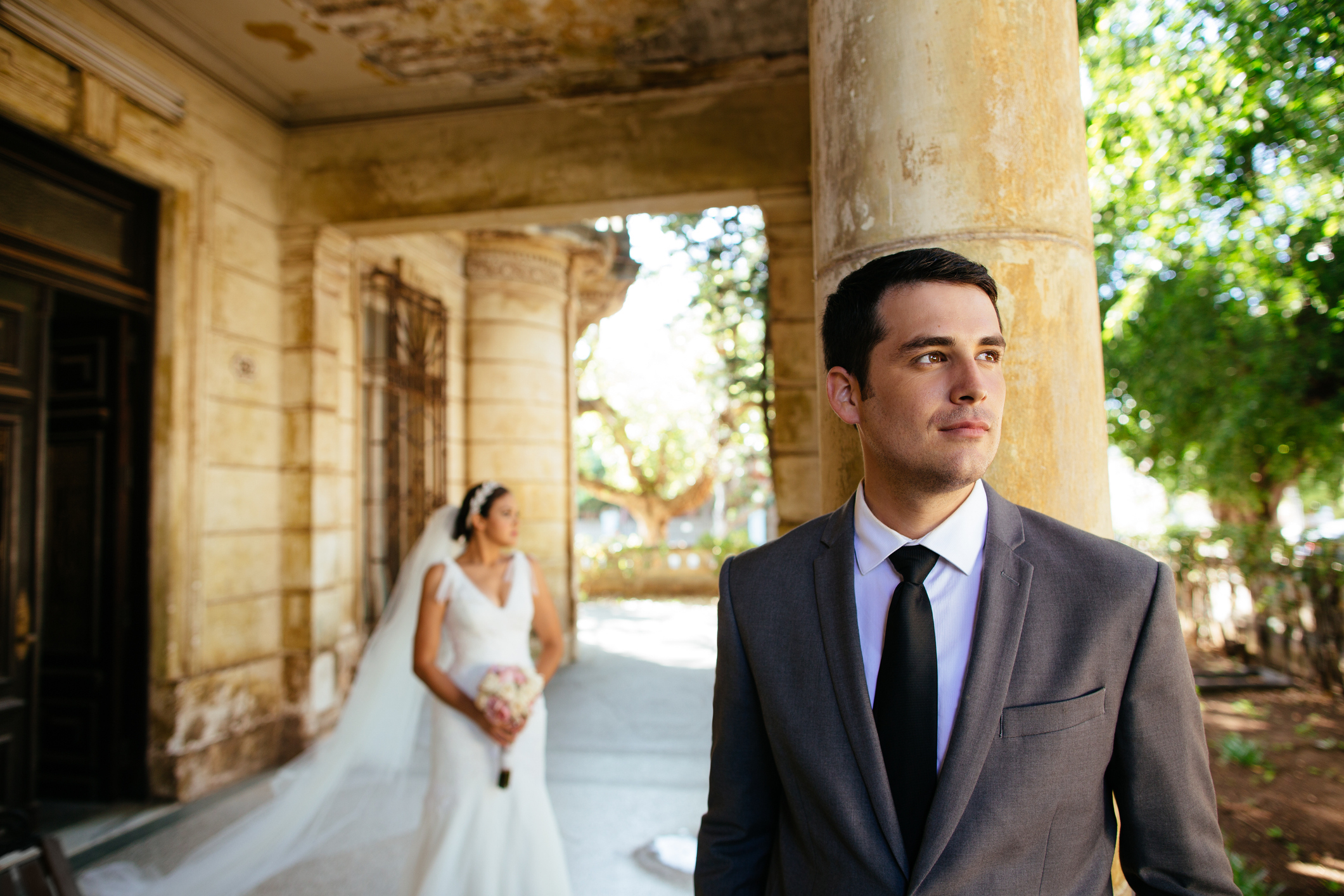 A groom in a suit and tie looks away, standing outside an old building. In the background, a bride in a wedding gown and veil holds a bouquet