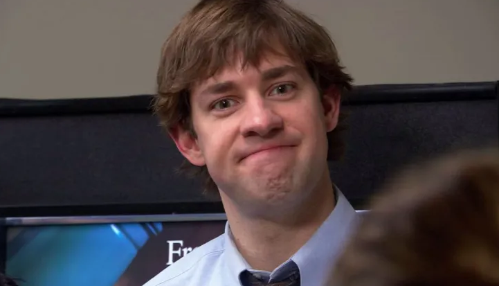 Man from a popular TV show standing indoors, looking towards the camera with a slight smile. The background is partially obscured by another person