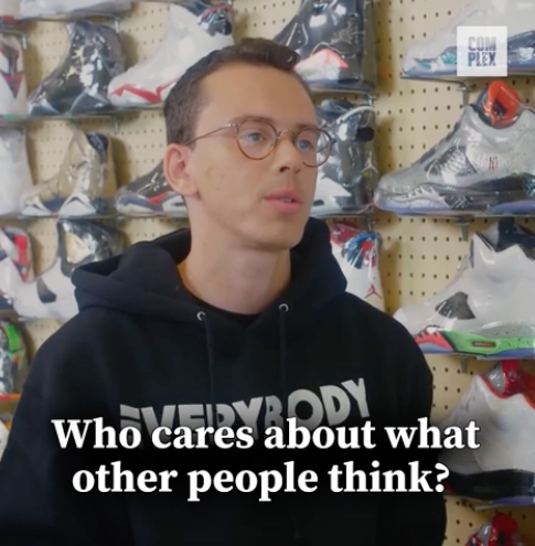 Logic stands in a sneaker store in a black hoodie with the text &quot;EVERYBODY.&quot; Caption below reads, &quot;Who cares about what other people think?&quot;