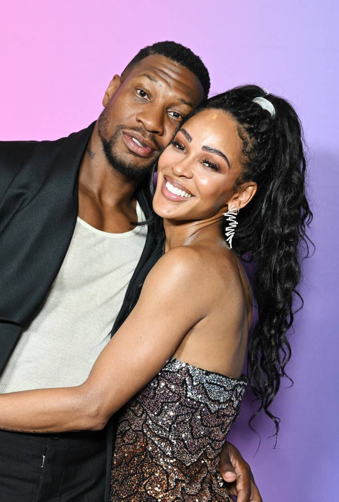 Jonathan Majors and Meagan Good hug and smile at an event. Majors wears a black jacket and white shirt; Good wears a strapless, shimmering dress