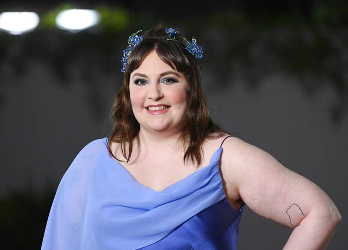 Lena Dunham smiles while posing on the red carpet, wearing a one-shoulder, flowing dress and a floral headband