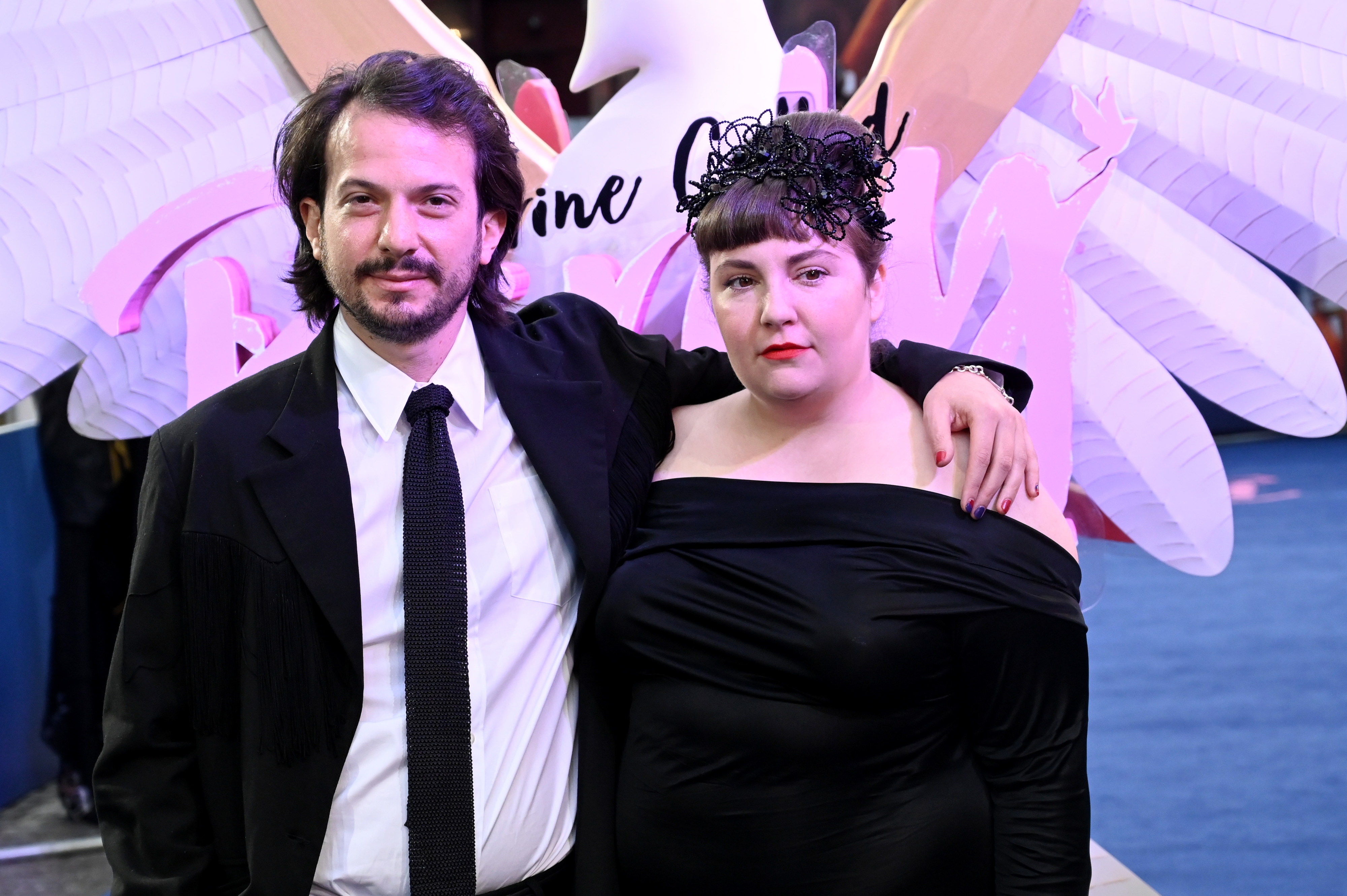 Lena Dunham and Luis Felber pose on a red carpet. Dunham wears an off-the-shoulder black dress and headpiece, and Felber wears a black suit and tie