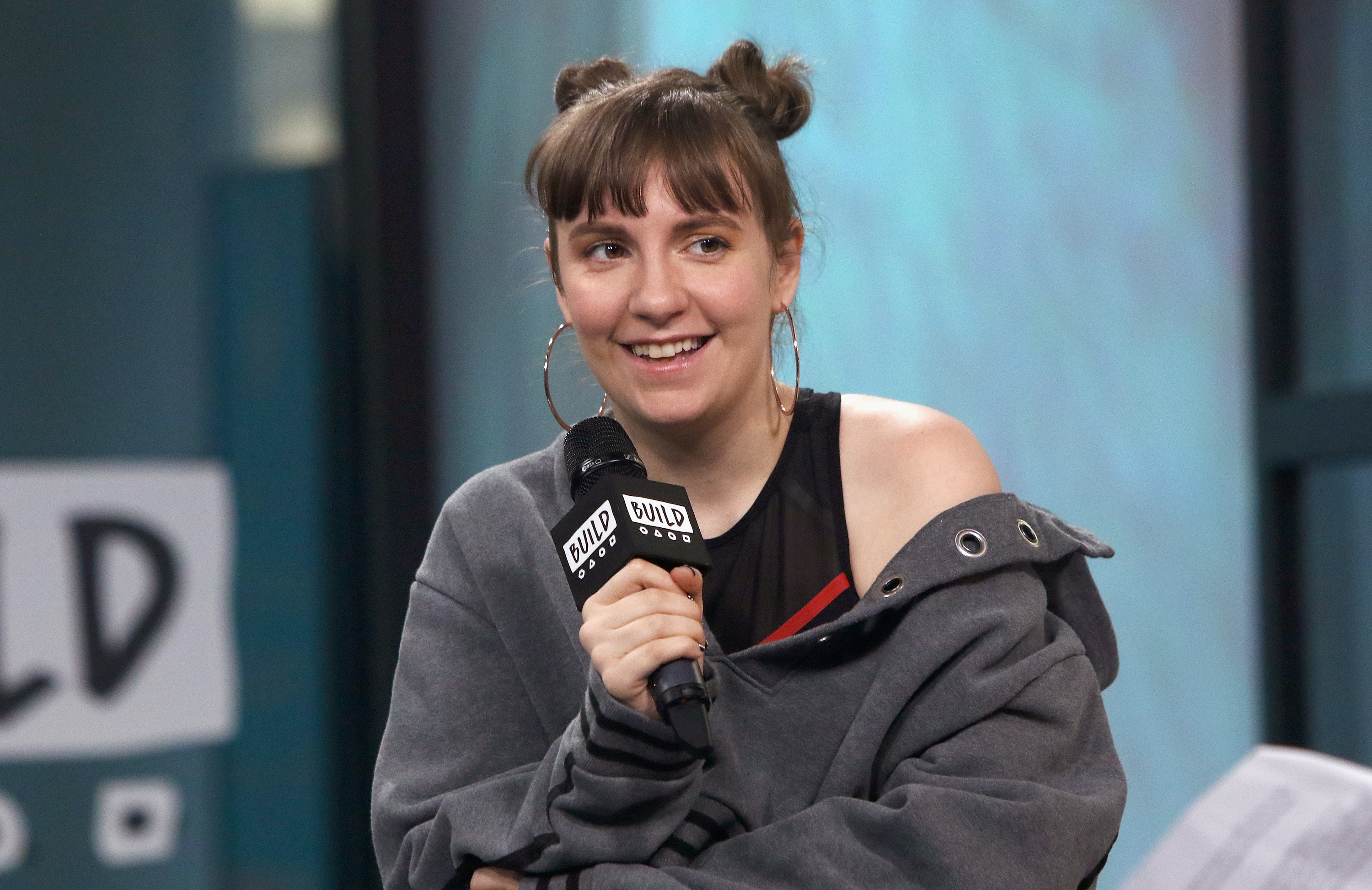 Lena Dunham wearing a casual outfit, with her hair in two buns, speaking into a microphone at a BUILD Series event