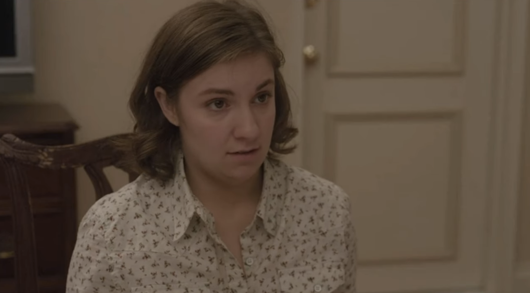 Lena Dunham sits in a room wearing a patterned shirt, looking slightly surprised. The background features a closed door and a wooden chair