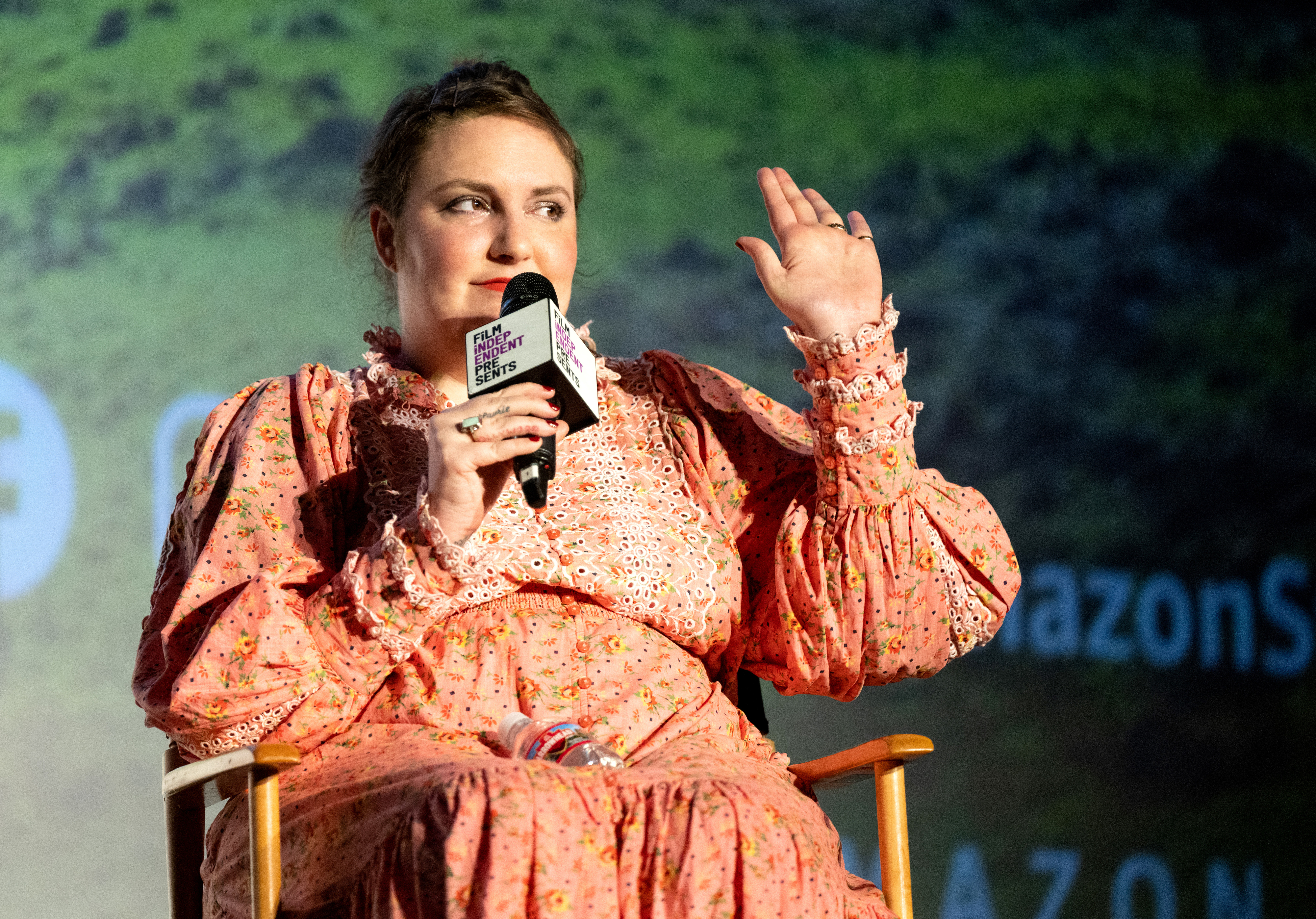 Lena Dunham speaks at an event, wearing a floral long-sleeve dress, holding a microphone, and gesturing with her other hand