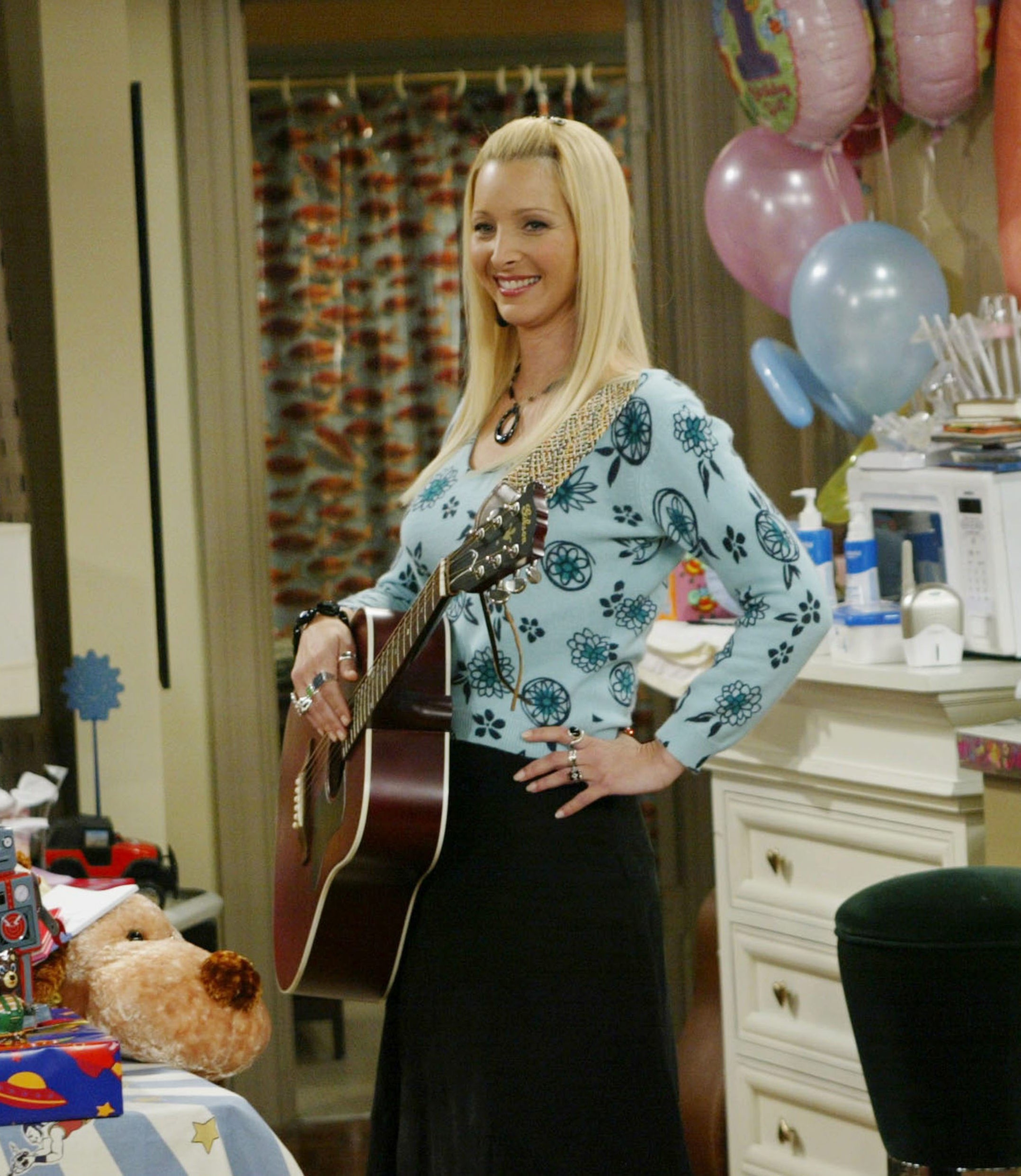 Lisa Kudrow as her character Phoebe Buffay in Friends stands at a children&#x27;s party holding a guitar, with gifts, balloons, and party decorations around her