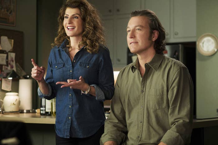 Nia Vardalos, in a denim shirt, gestures while standing next to John Corbett, in a button-up shirt. They are in a kitchen setting