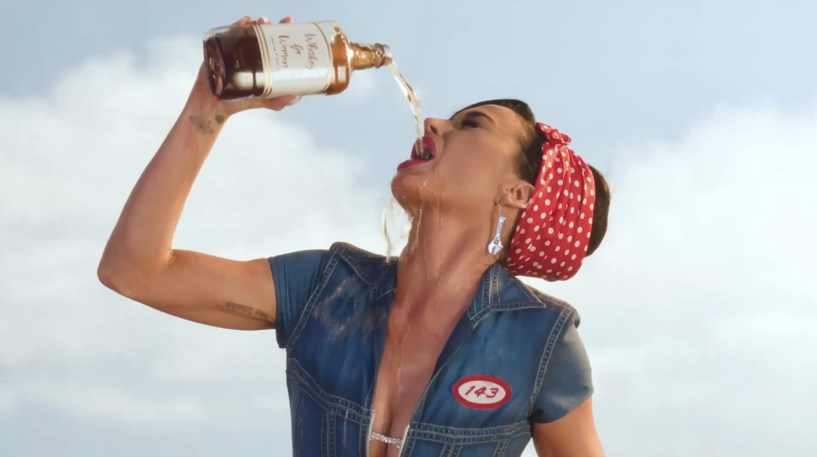 A person with a red polka dot headband and denim attire is pouring a bottle of liquid into their mouth with the liquid spilling