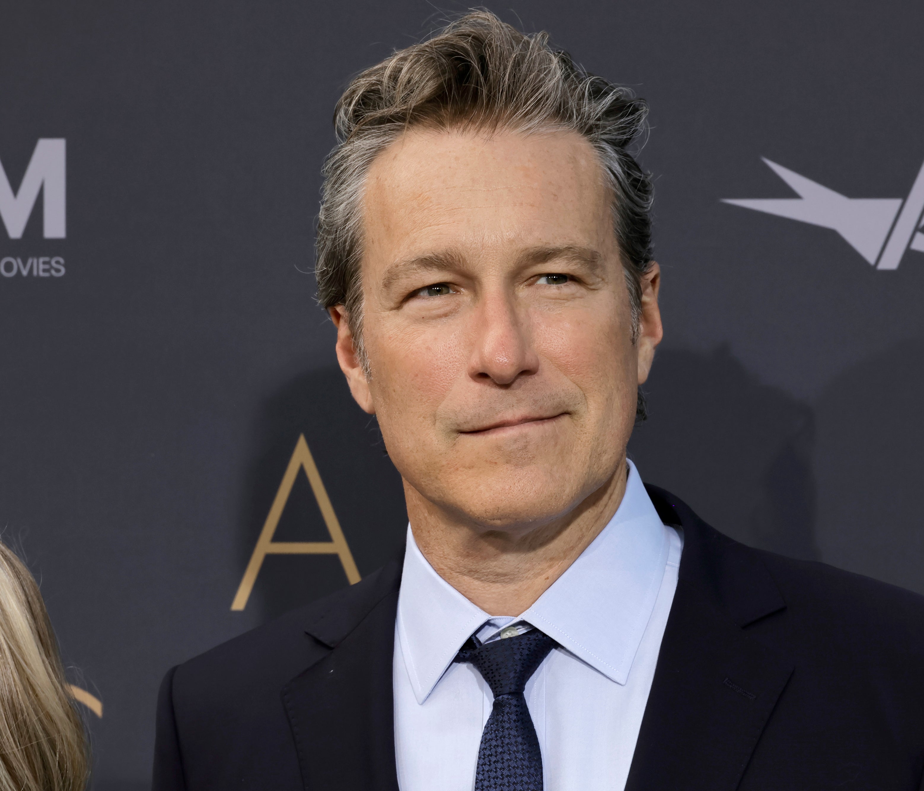 John Corbett, wearing a suit and tie, stands on a red carpet at an event