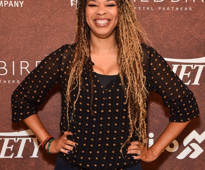 Nkechi smiles broadly, standing with hands on hips, wearing a black blouse and dark jeans, at a Variety-sponsored event