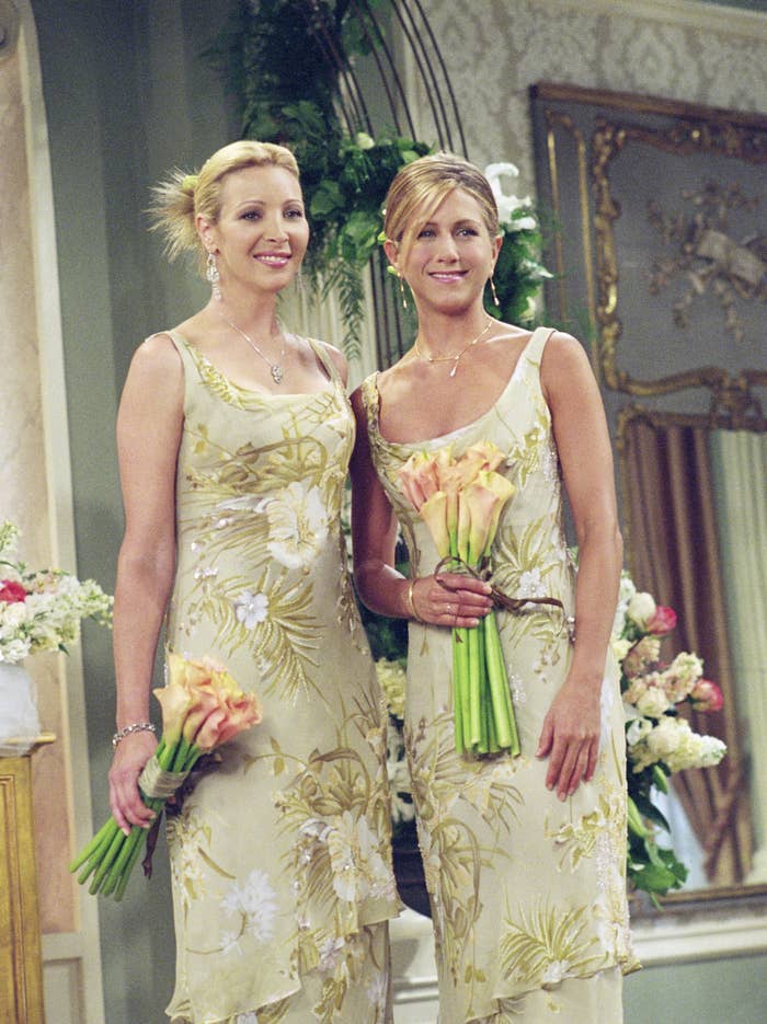 Lisa Kudrow and Jennifer Aniston are standing side by side, smiling and holding bouquets, both wearing floral-patterned dresses in an elegant setting