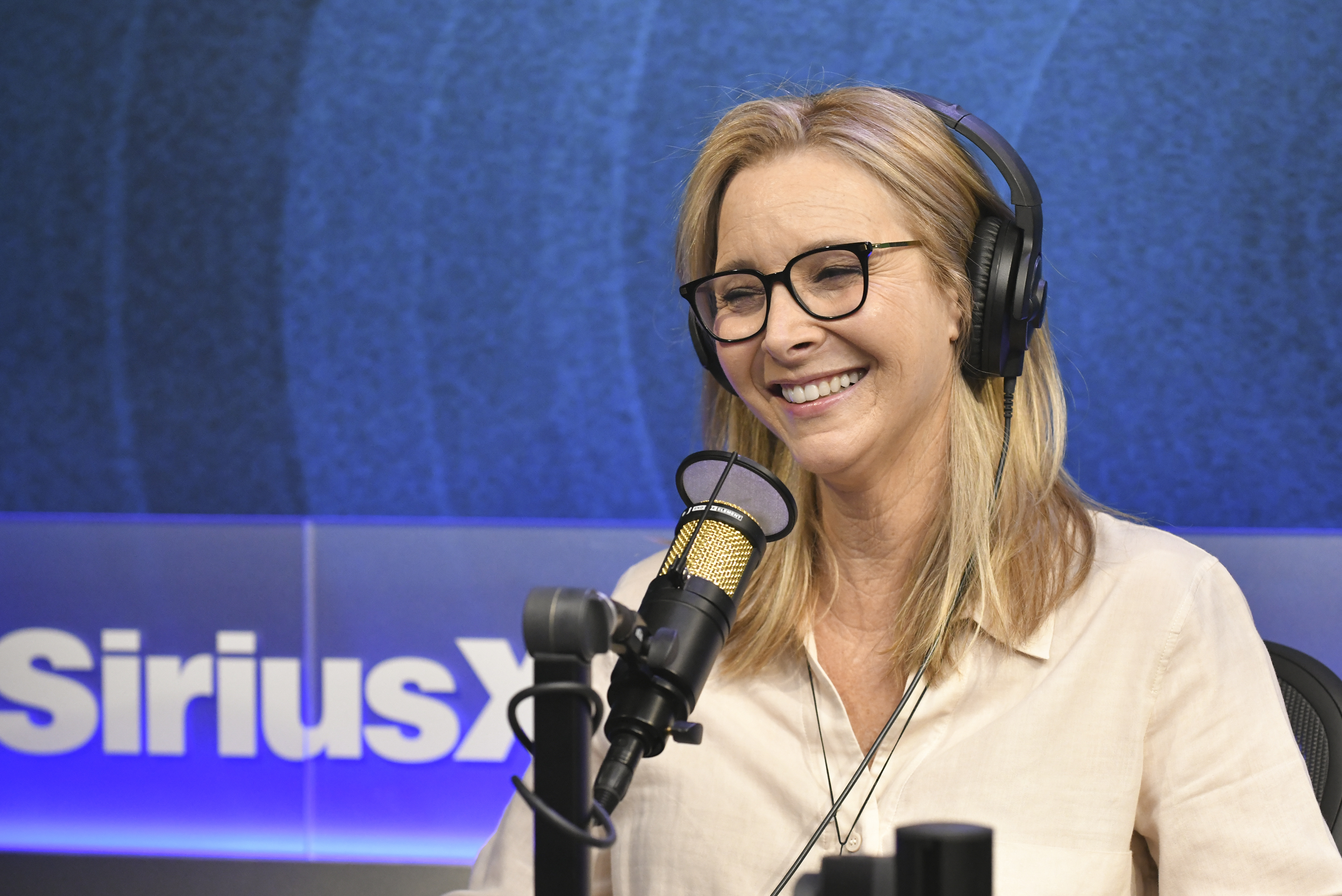 Lisa Kudrow smiling and speaking during a radio interview at SiriusXM studios, wearing a casual blouse, glasses, and headphones