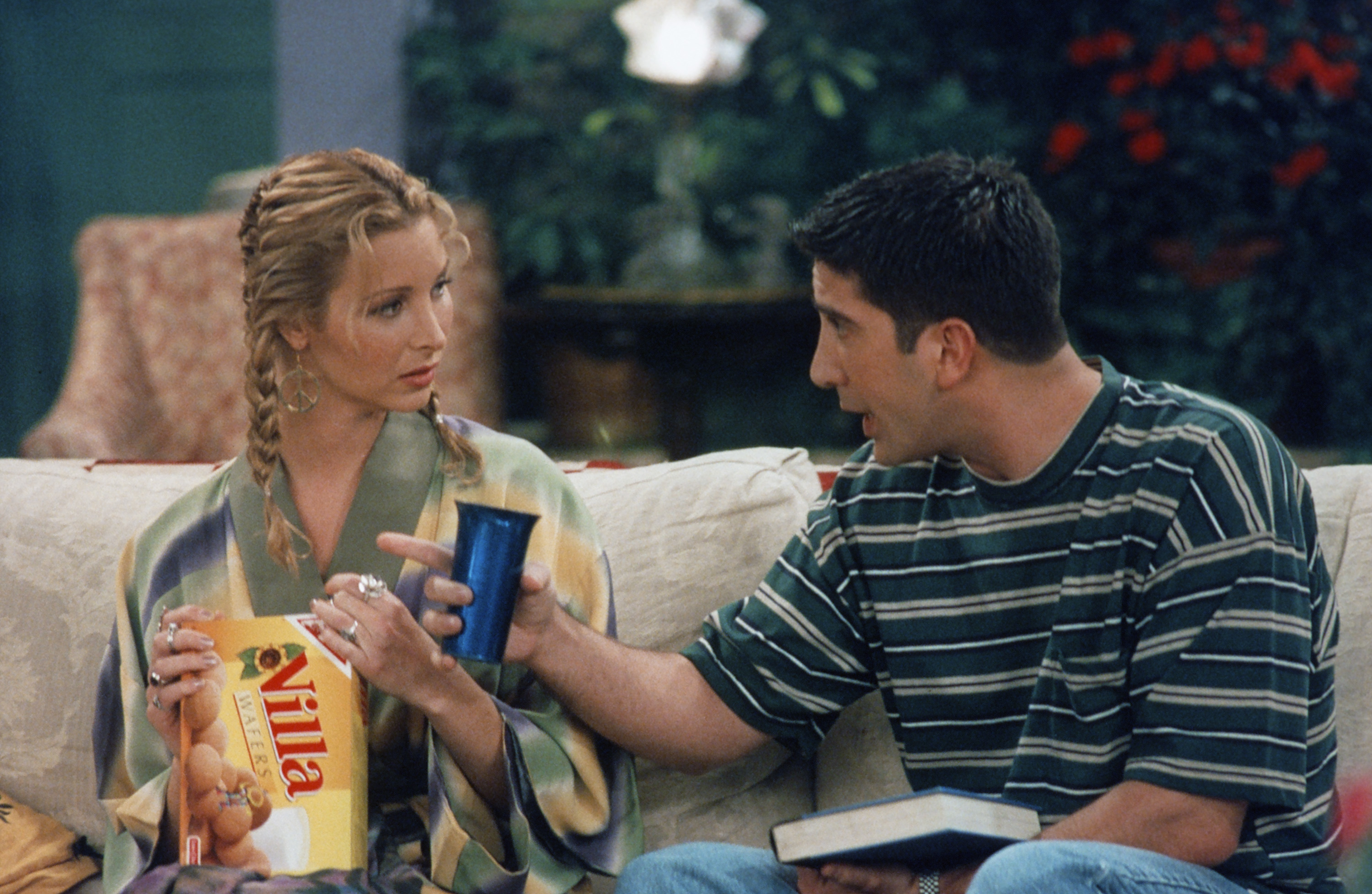 Lisa Kudrow, in a robe, holds a box of Vanilla Wafers while David Schwimmer, in a striped shirt, gestures to her with a cup on a sofa