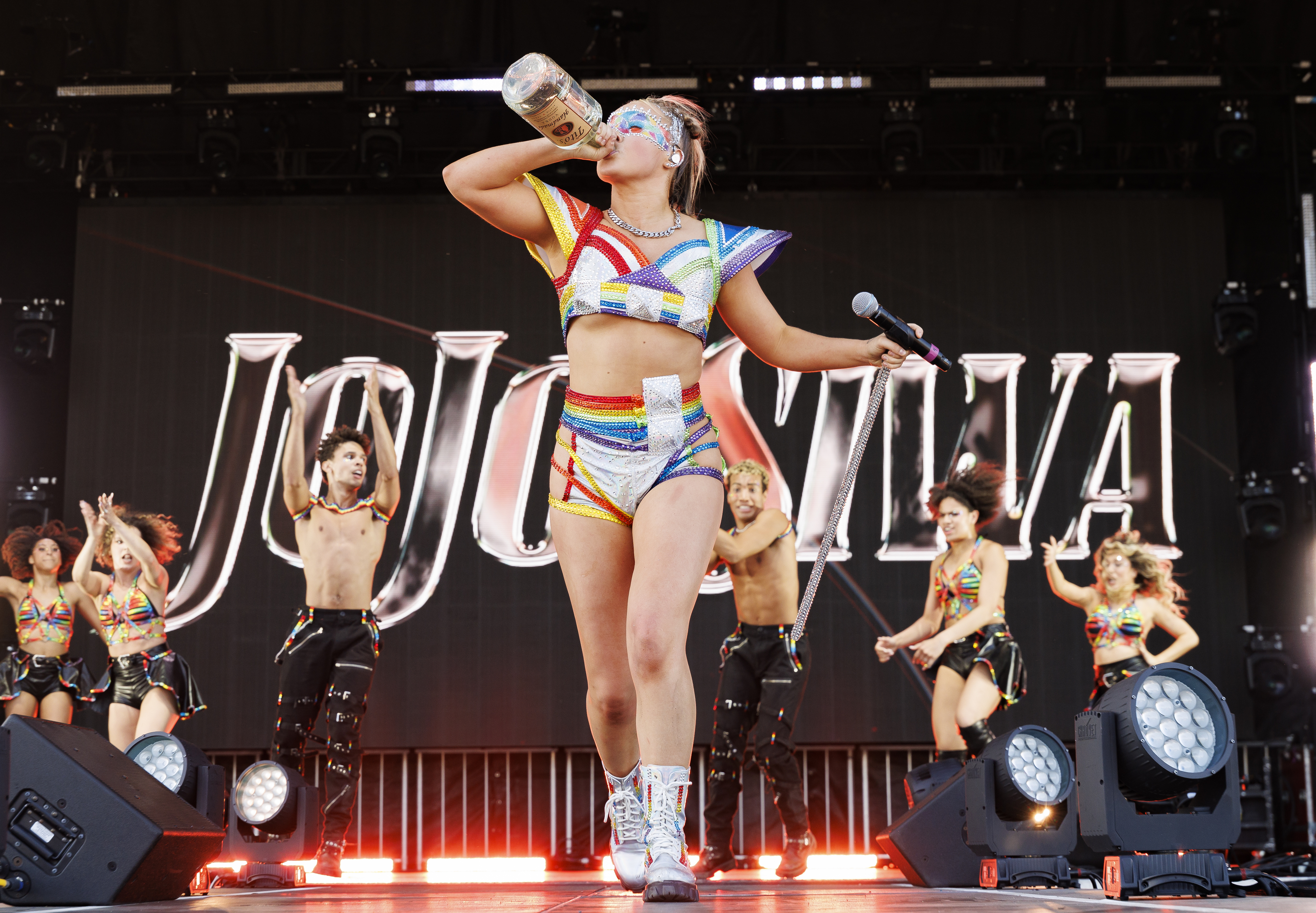 JoJo Siwa performs on stage in a rainbow-themed outfit with backup dancers in similar outfits. She is seen drinking from a large bottle while holding a microphone