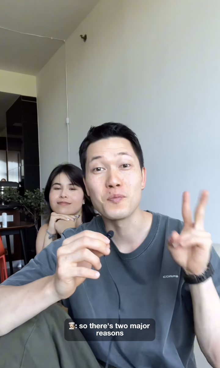 A man and woman sit casually indoors. The man speaks into a microphone and gestures with two fingers. Subtitles say: &quot;so there&#x27;s two major reasons&quot;