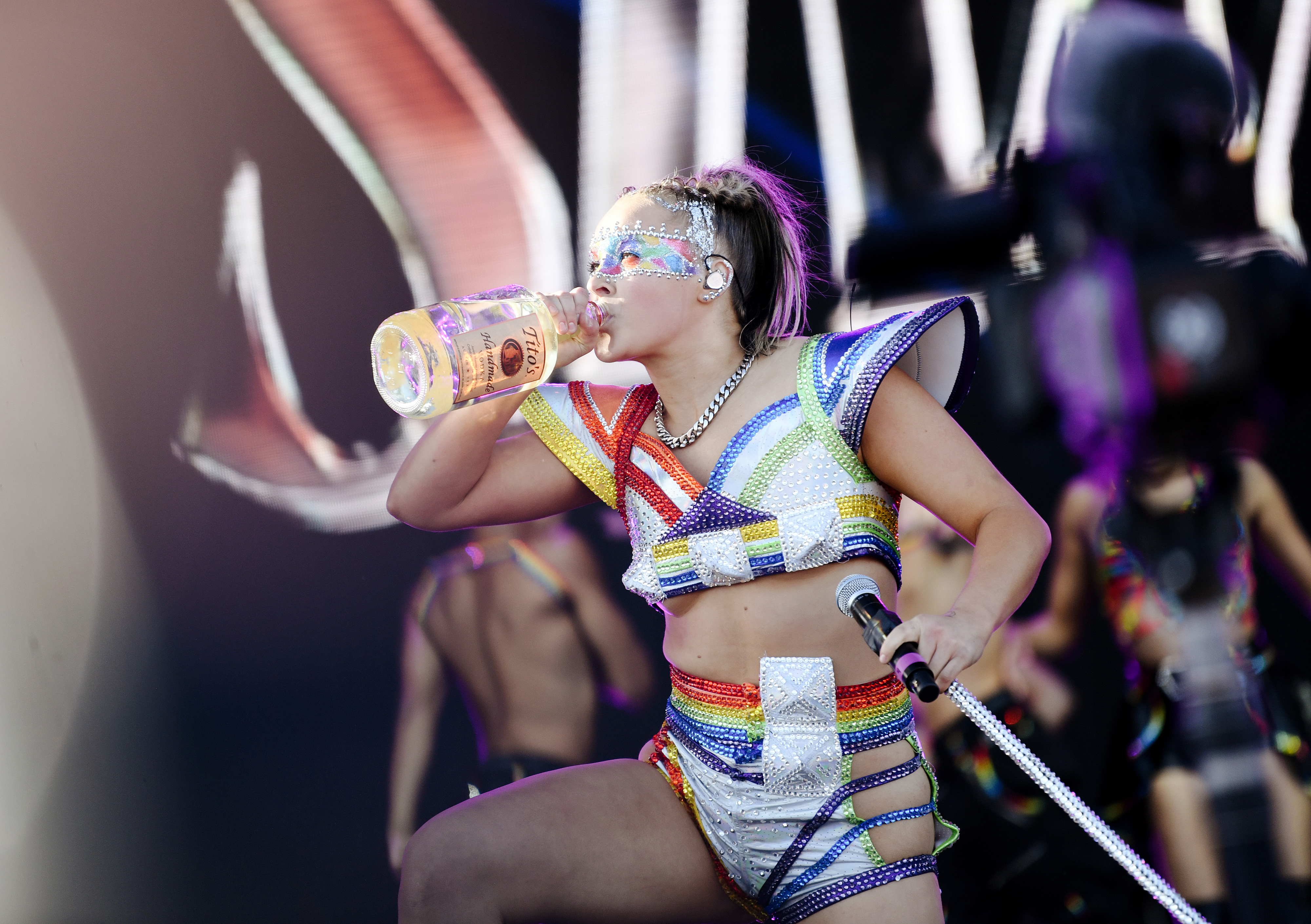 Jojo Siwa performs on stage, wearing an elaborate outfit with geometric shapes and sparkles, while drinking from a large bottle