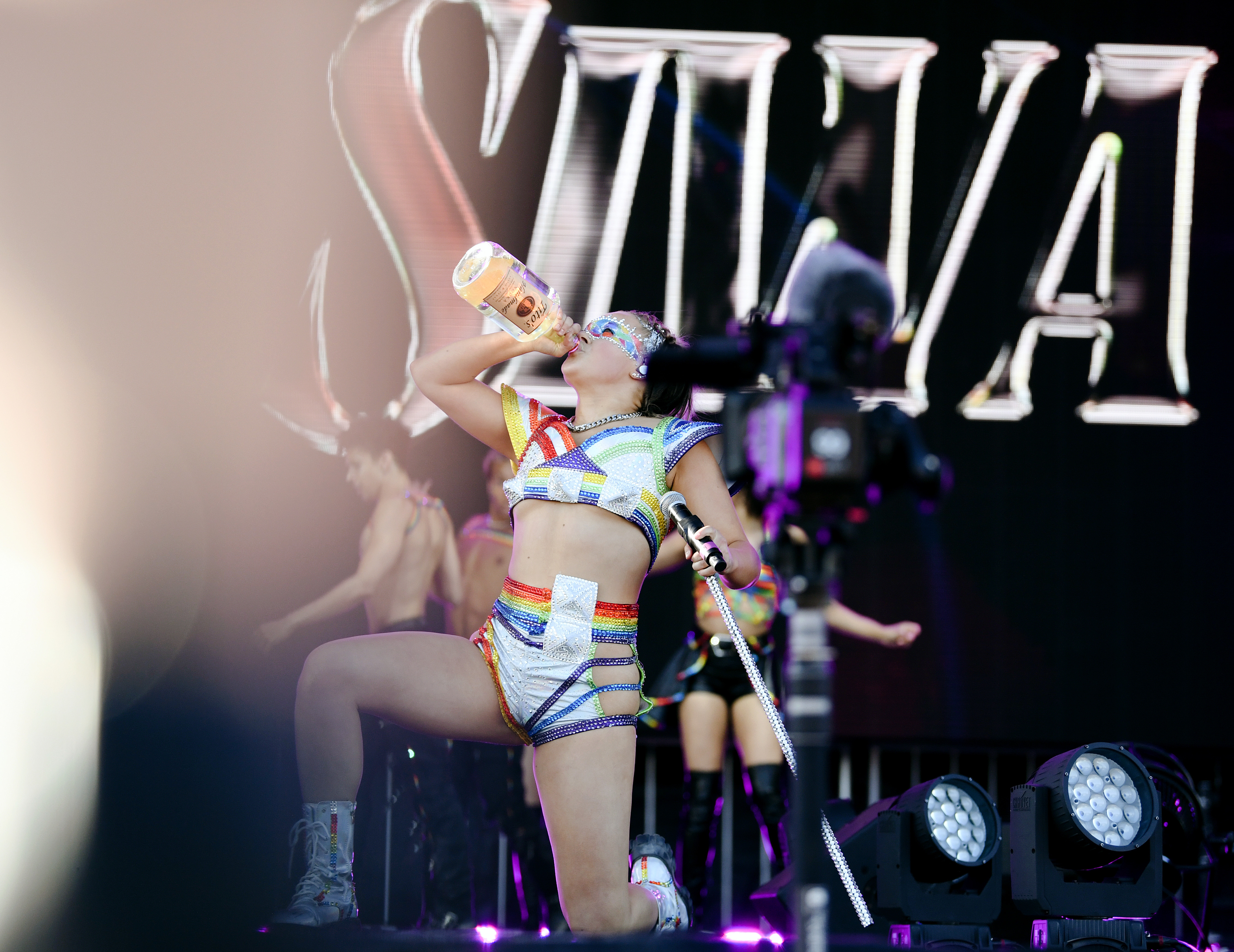 Singer JoJo Siwa performs on stage wearing a futuristic, multi-colored outfit, drinking from a large bottle, with backup dancers in the background