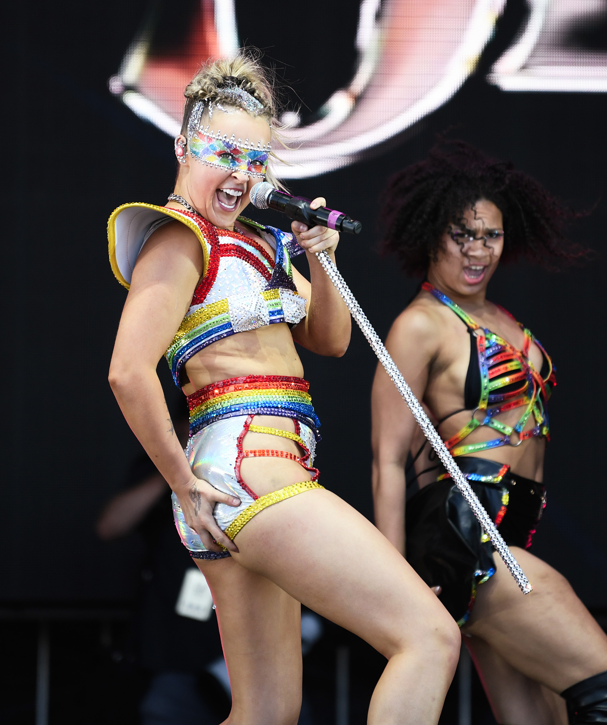 JoJo Siwa and a dancer perform on stage. JoJo wears a rainbow-themed, sequin-studded outfit with a matching visor, and holds a sparkling mic stand