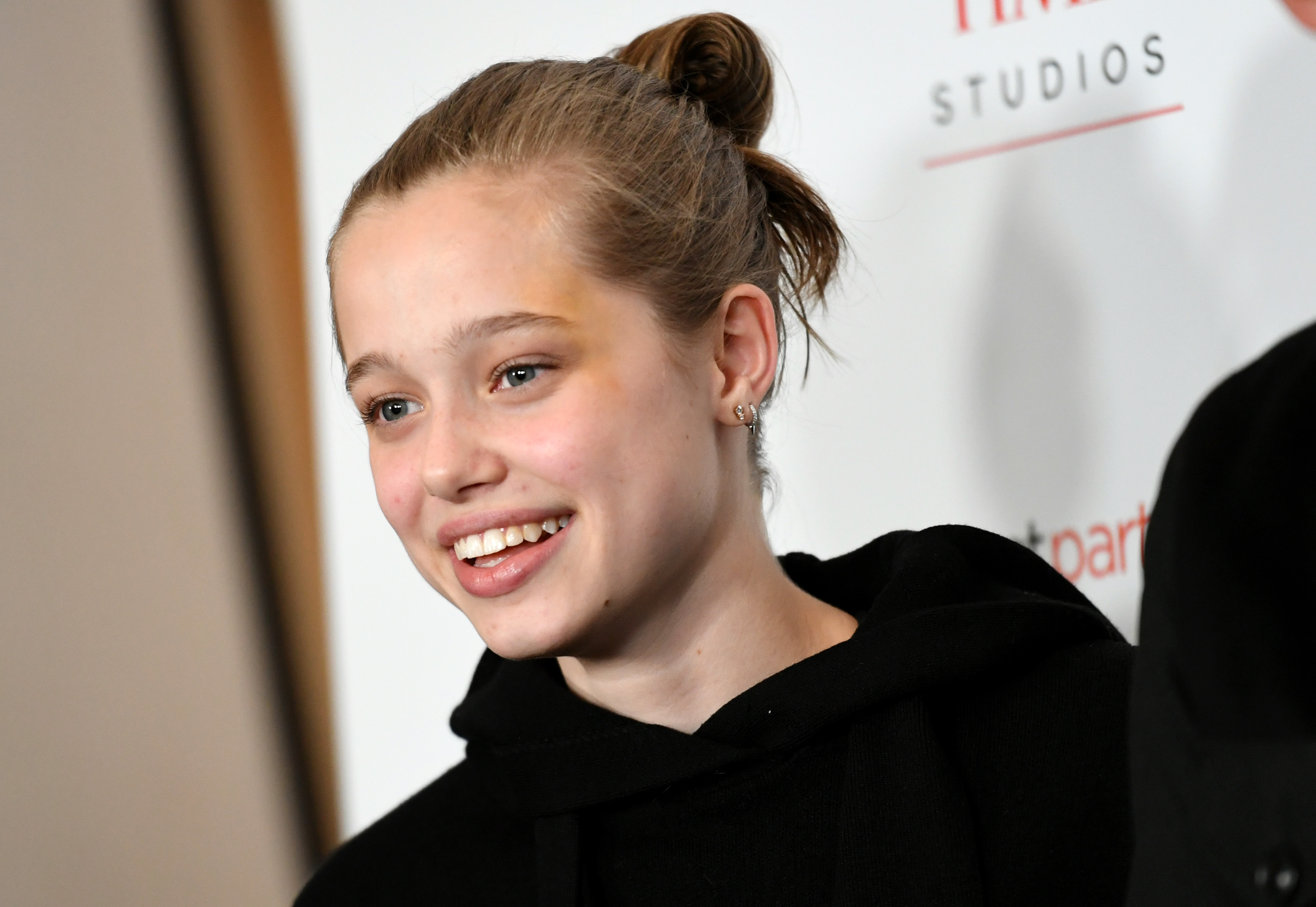 Shiloh Jolie smiles at a red carpet event, wearing a casual black hoodie with hair styled in a bun