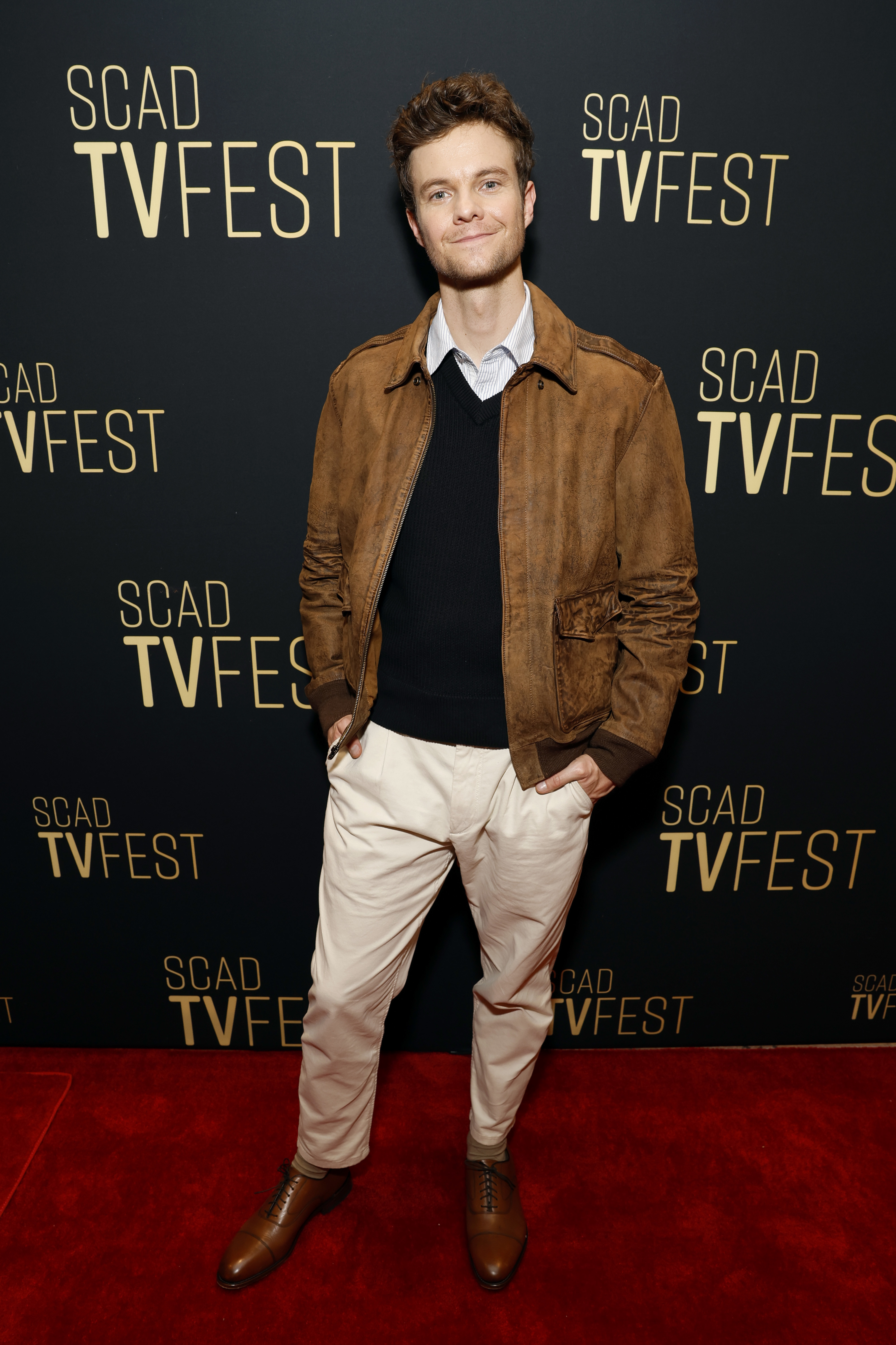 Jack Quaid poses on the red carpet at SCAD TVfest, wearing a brown jacket over a black sweater and white pants
