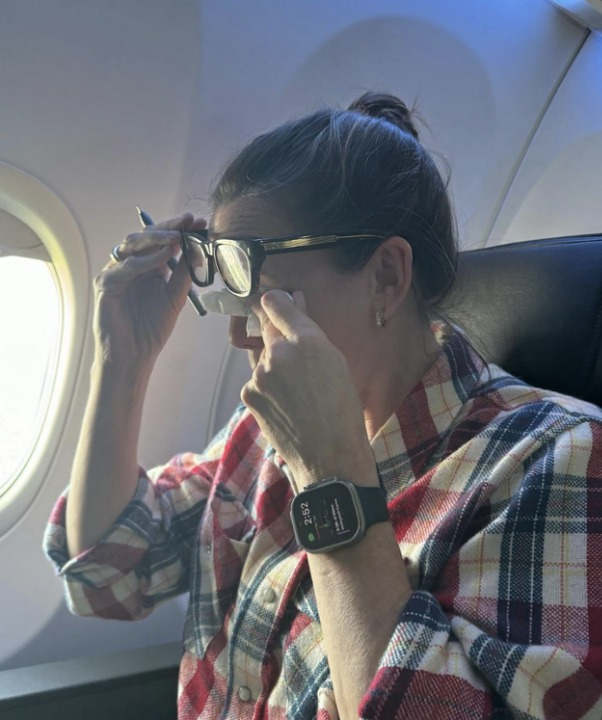 A woman on an airplane wearing a plaid shirt is adjusting her glasses while seated next to an airplane window. She also has an Apple Watch on her wrist