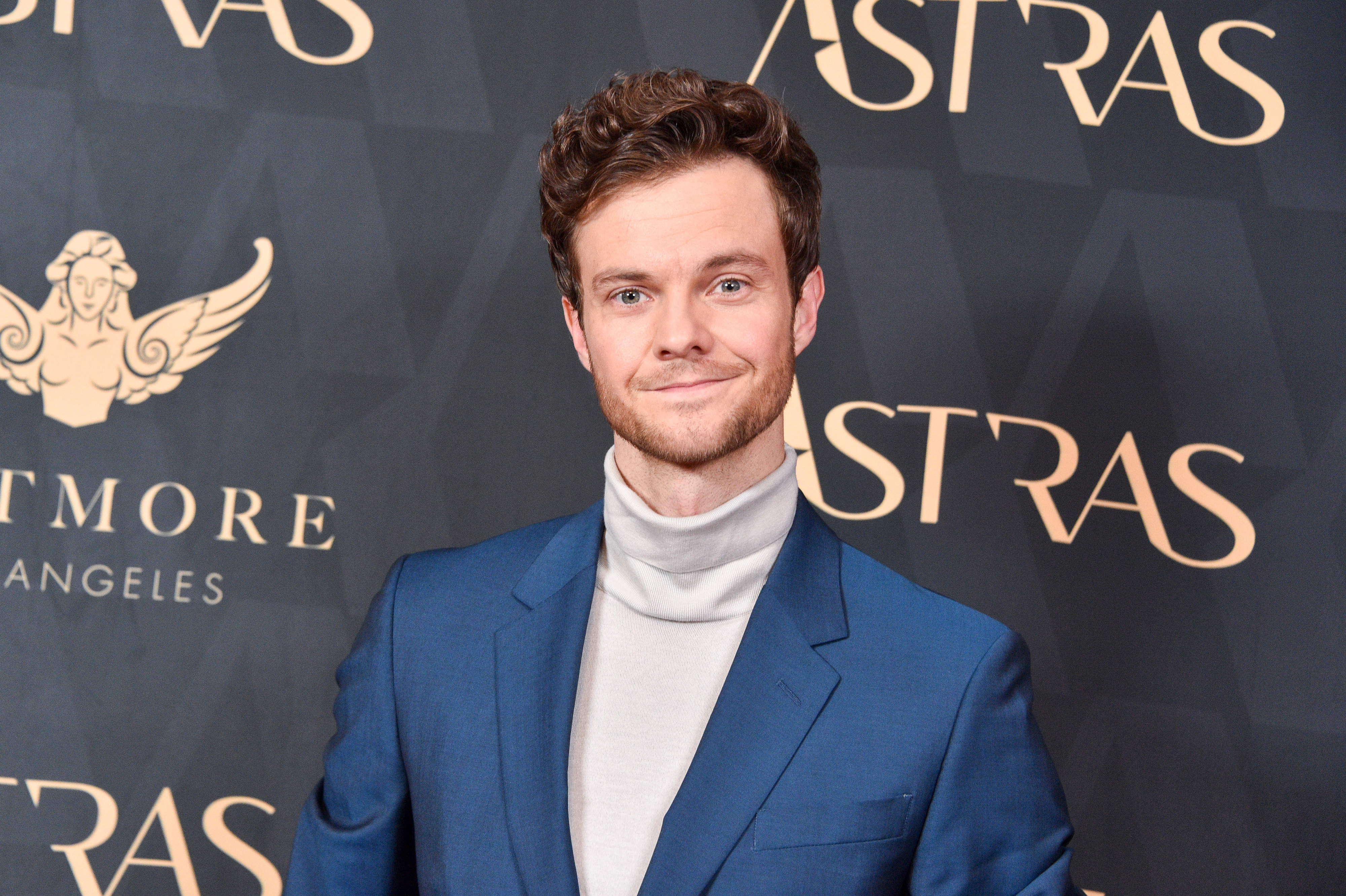 Jack Quaid, in a blue suit with a white turtleneck, poses on the red carpet at an event